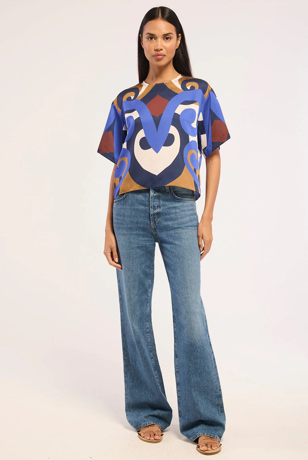 A woman modeling a short-sleeve crop top with a bold abstract pattern in blue, brown, beige, and white, paired with wide-leg blue jeans and tan sandals, against a plain light background.