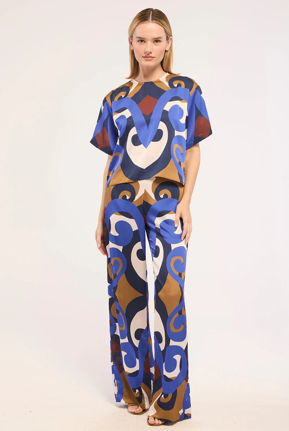 A woman modeling a matching short-sleeve top and wide-leg pants set with a bold abstract pattern in blue, brown, beige, and white, photographed against a plain light background.