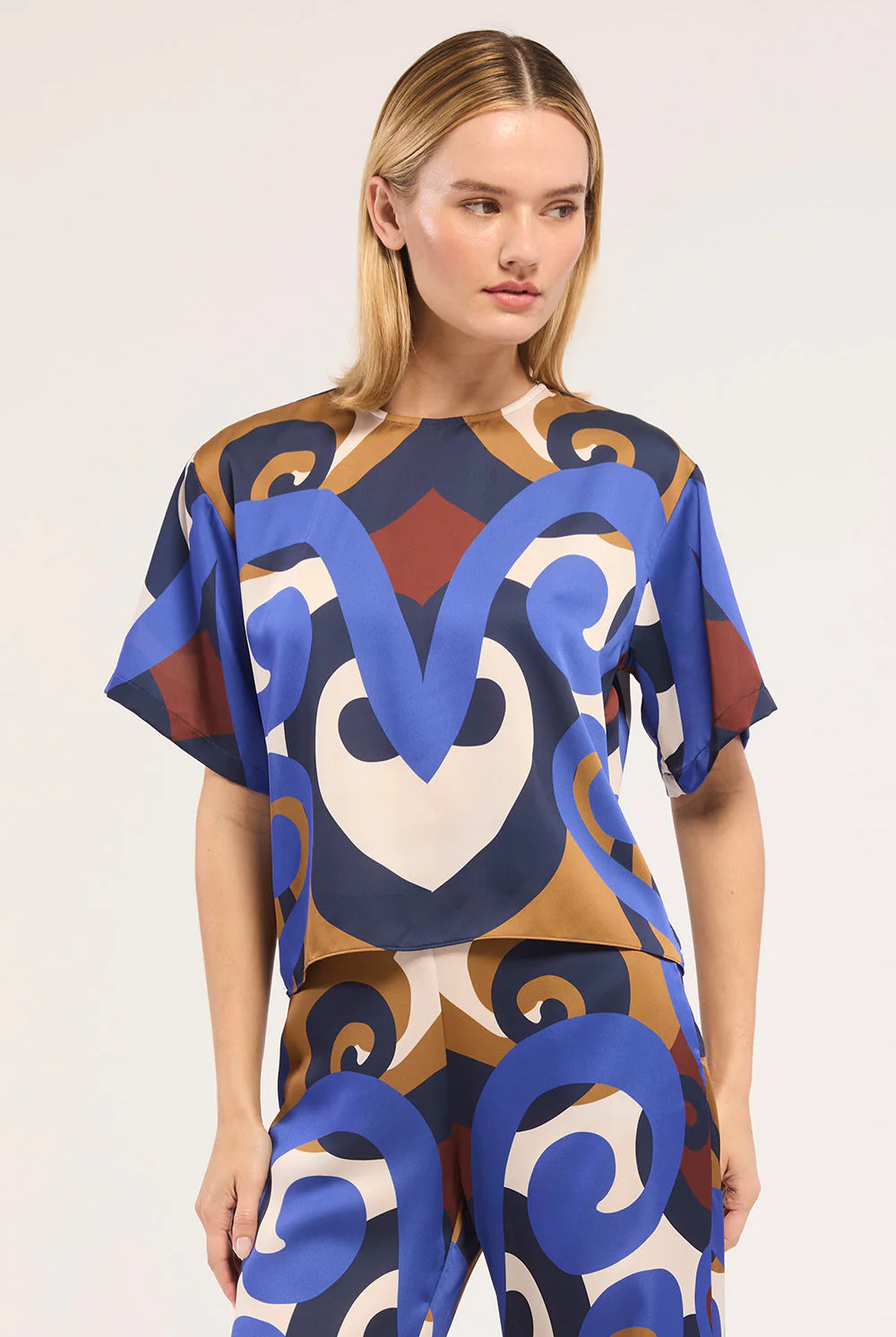A woman modeling a matching short-sleeve top and pants set with a bold abstract pattern in blue, tan, brown, and cream against a plain light background.