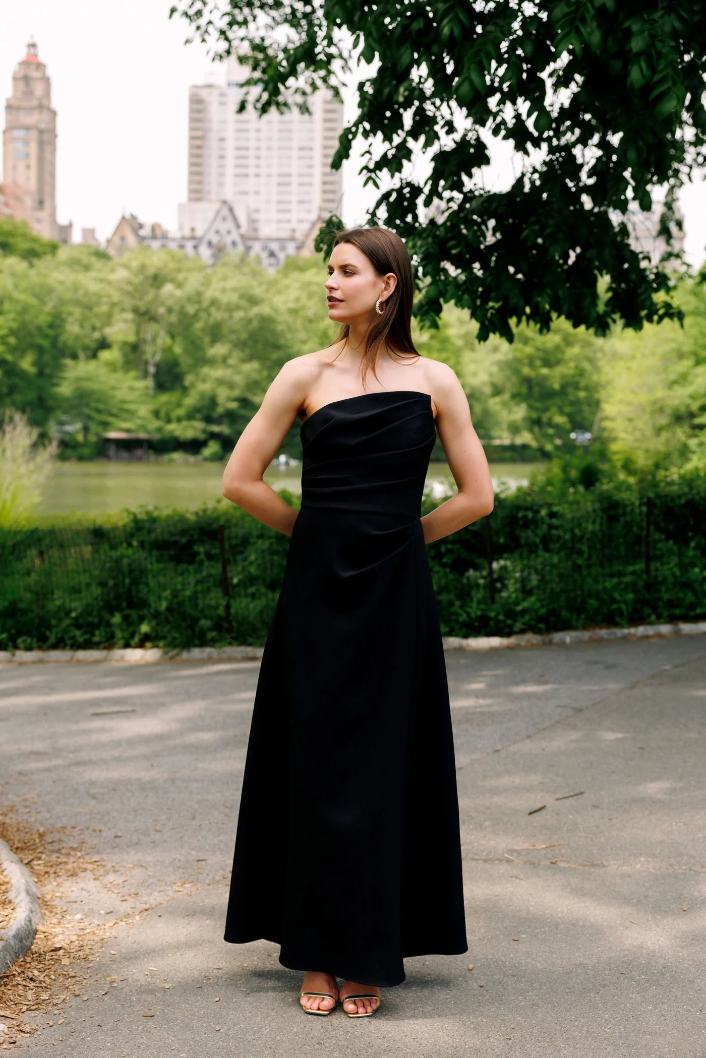 A woman wearing a strapless black floor-length gown stands outdoors on a paved path in a park, with trees, greenery, and buildings in the background.