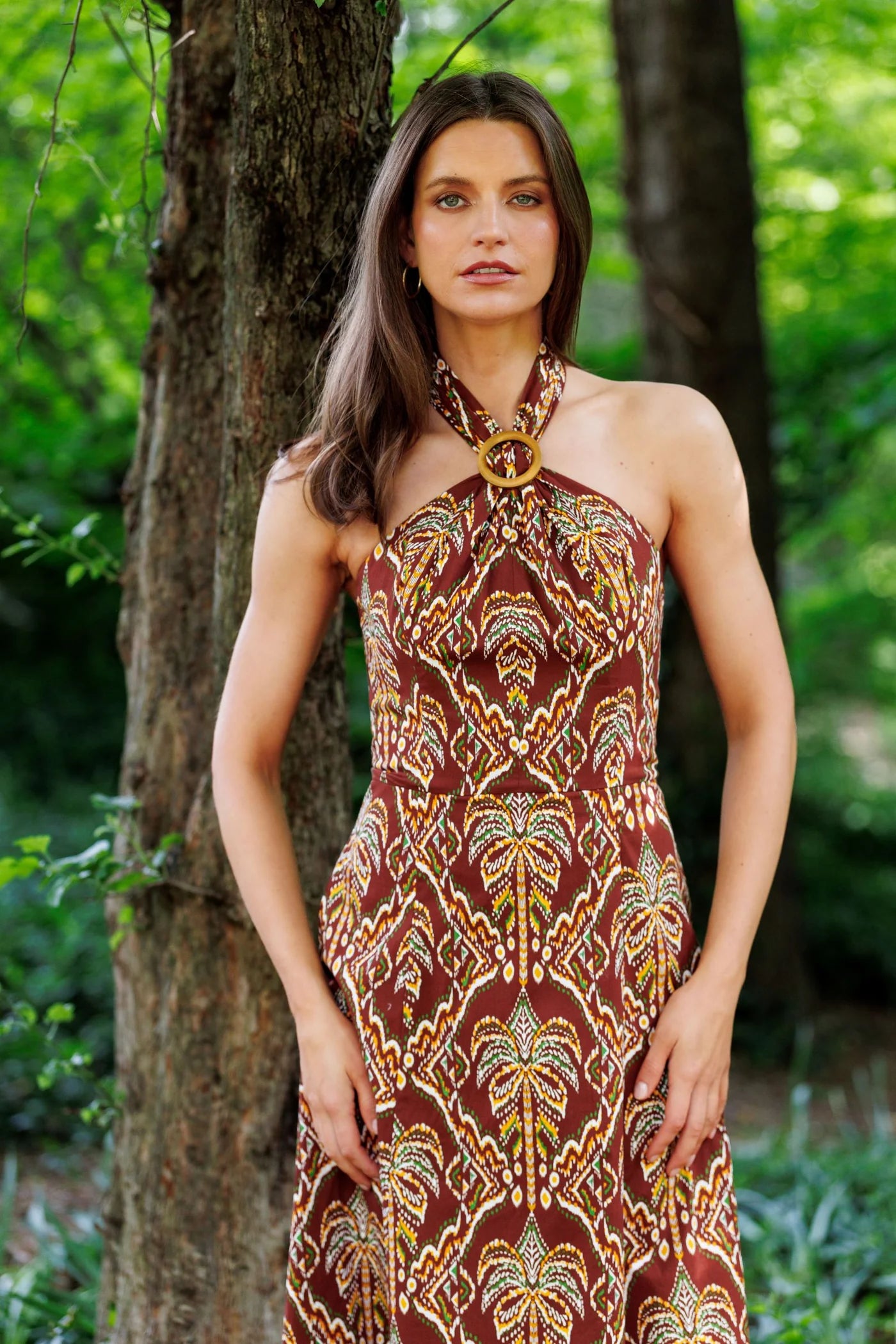 A woman wearing a brown halter-neck maxi dress with a gold and beige geometric pattern, standing outdoors in a wooded area.