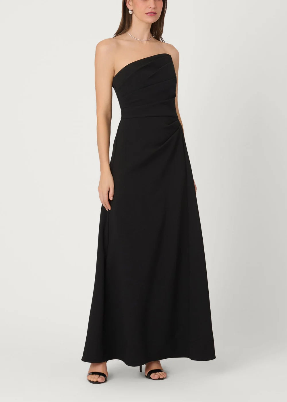 A woman modeling a strapless black maxi dress with a ruched bodice and flowing skirt, paired with black heeled sandals, against a light gray studio background.