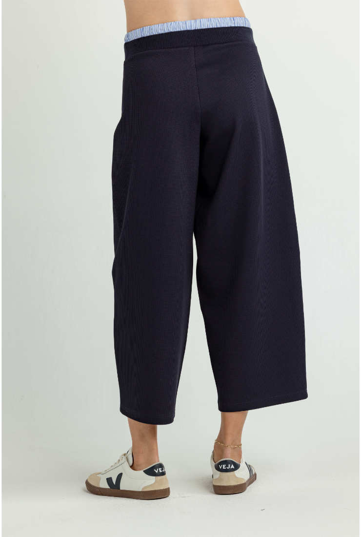 A model is shown from the back wearing dark navy wide-leg cropped pants with a light blue and white striped waistband, paired with beige and navy sneakers, against a plain light gray studio background.