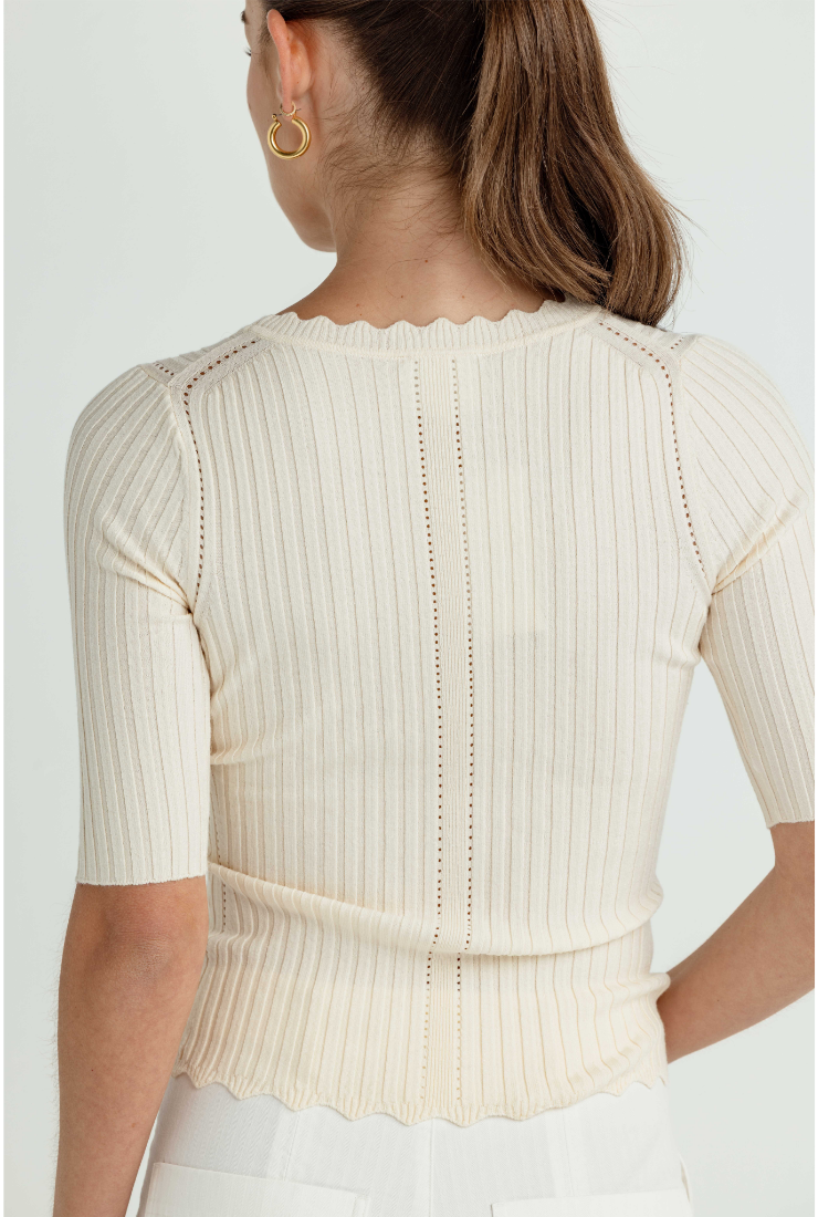 A woman shown from the back wearing a cream-colored ribbed short-sleeve top with a scalloped neckline and hem, paired with white bottoms against a light gray studio background.