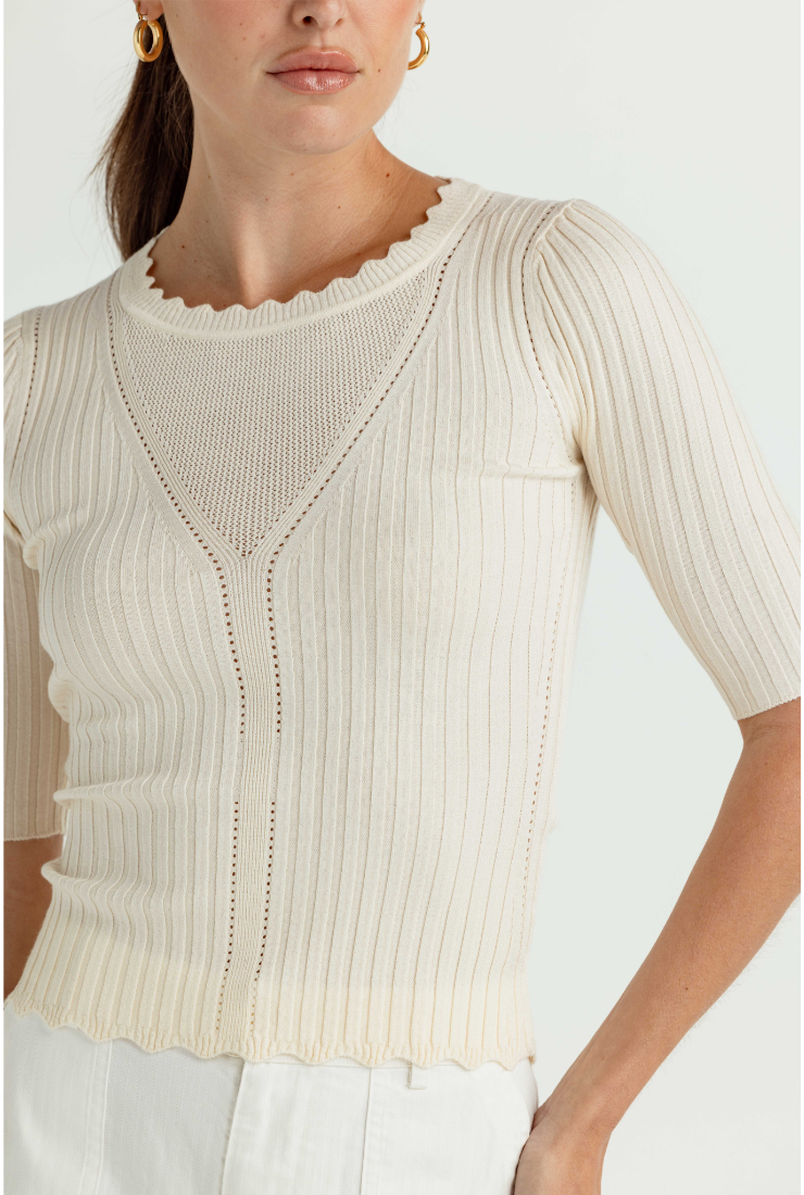 A close-up of a model wearing a ribbed knit short-sleeve top in cream with a scalloped neckline and front seam detailing, paired with white pants against a light gray studio background.