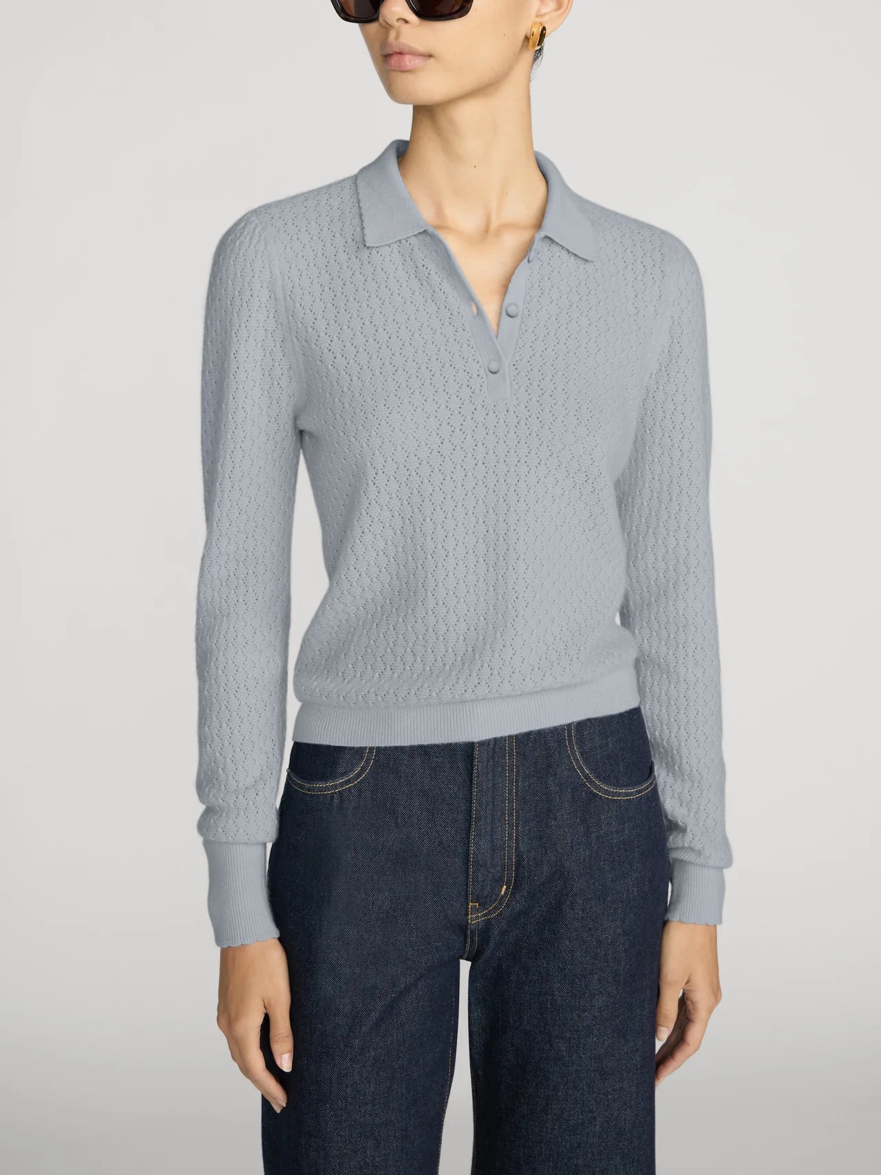 A model wearing a light gray textured knit polo sweater with a buttoned collar, paired with dark blue jeans, against a plain light gray studio background.