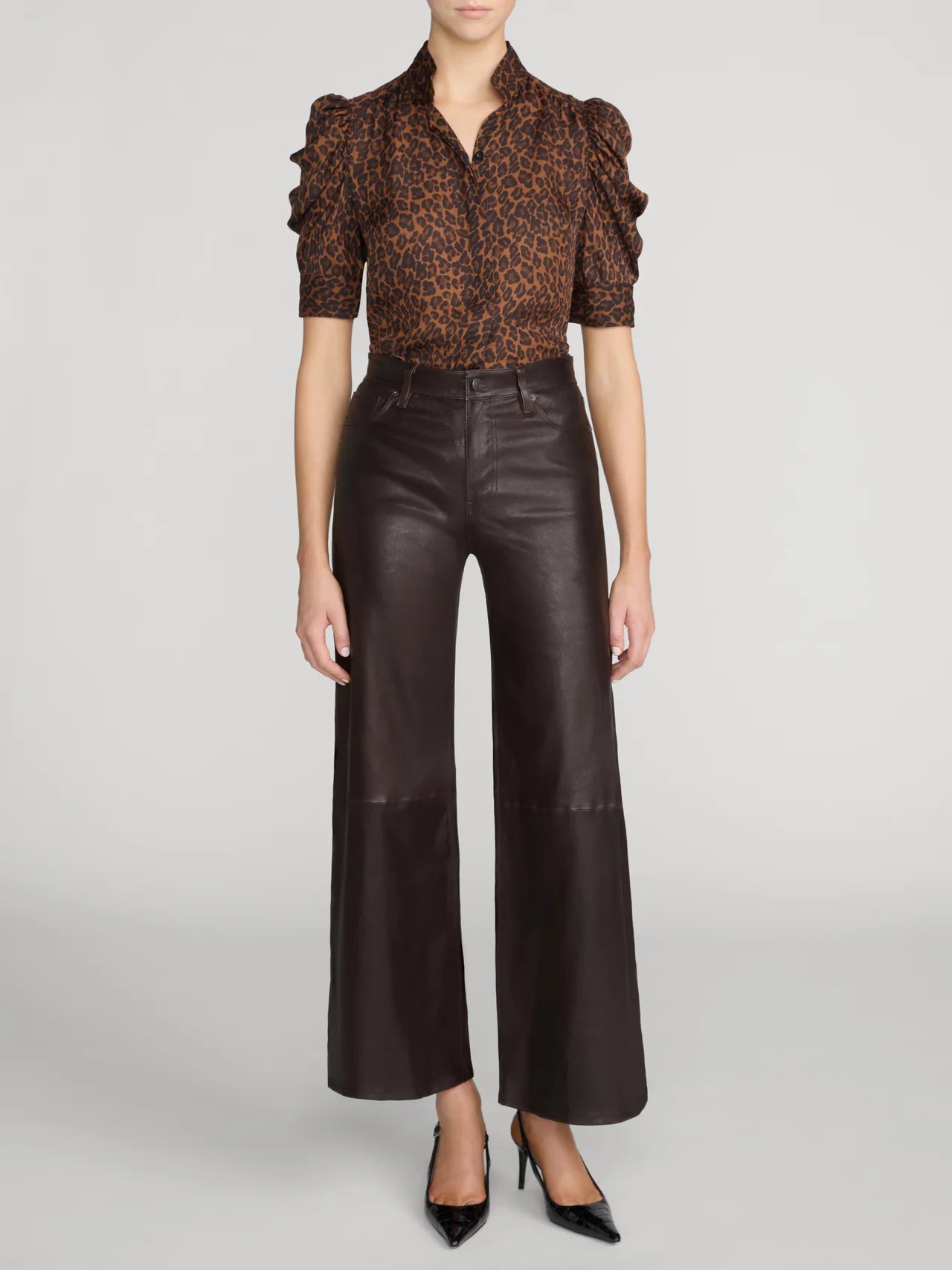 A model wearing a brown and black leopard-print short-sleeve blouse with ruffled sleeves, paired with dark brown wide-leg faux-leather pants and black pointed-toe heels, against a light gray studio background.