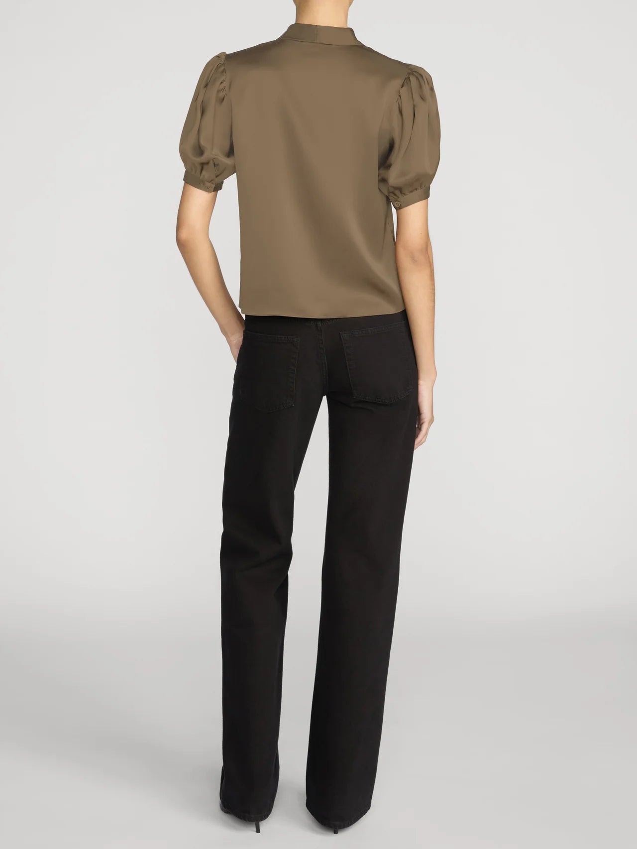 A model shown from the back wearing a short-sleeve olive brown blouse with puffed sleeves and black trousers, set against a plain light gray studio background.