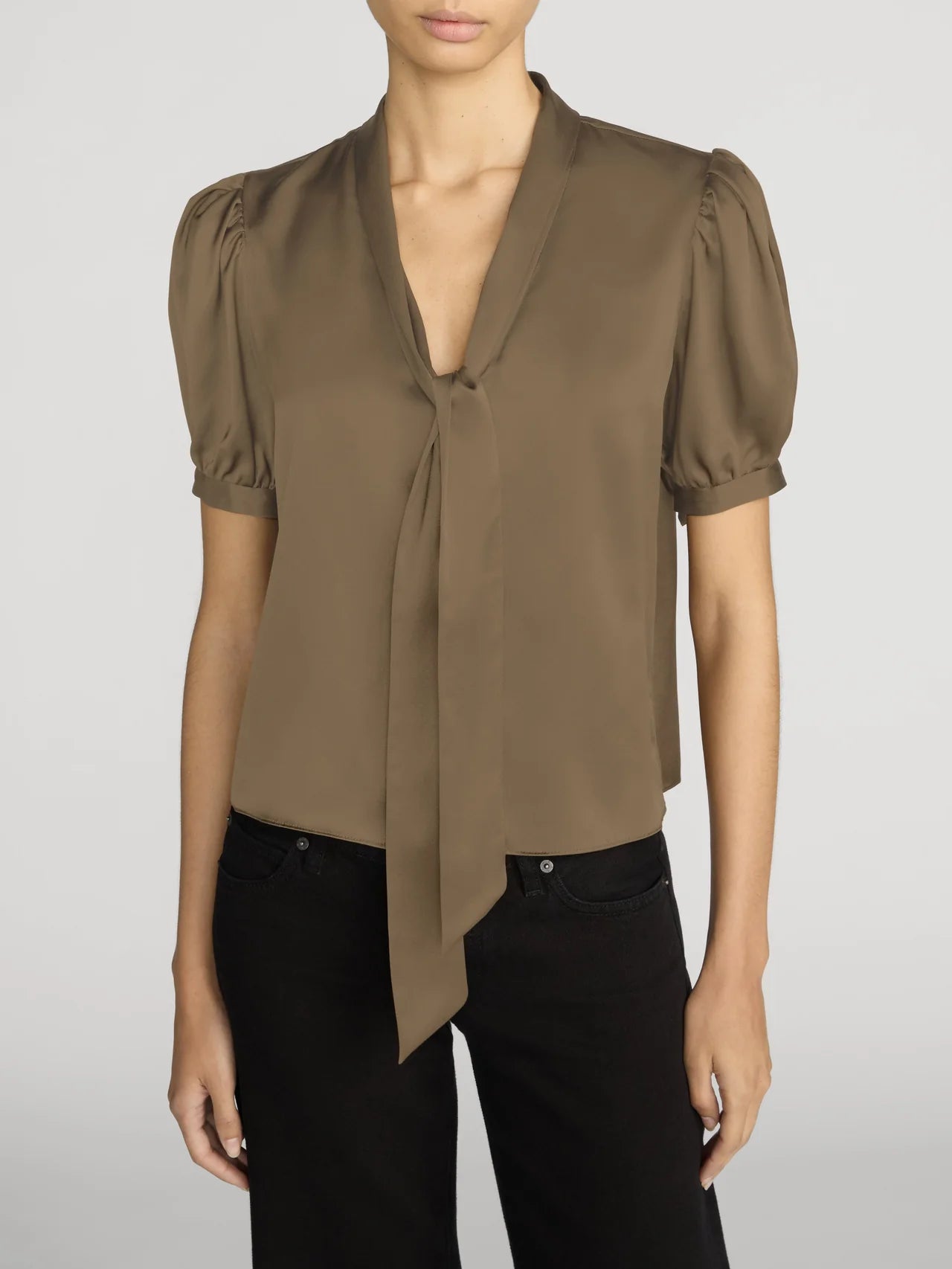 A woman wearing a taupe brown short-sleeve blouse with puff sleeves, a V-neckline, and a front tie detail, paired with black pants, against a light gray studio background.