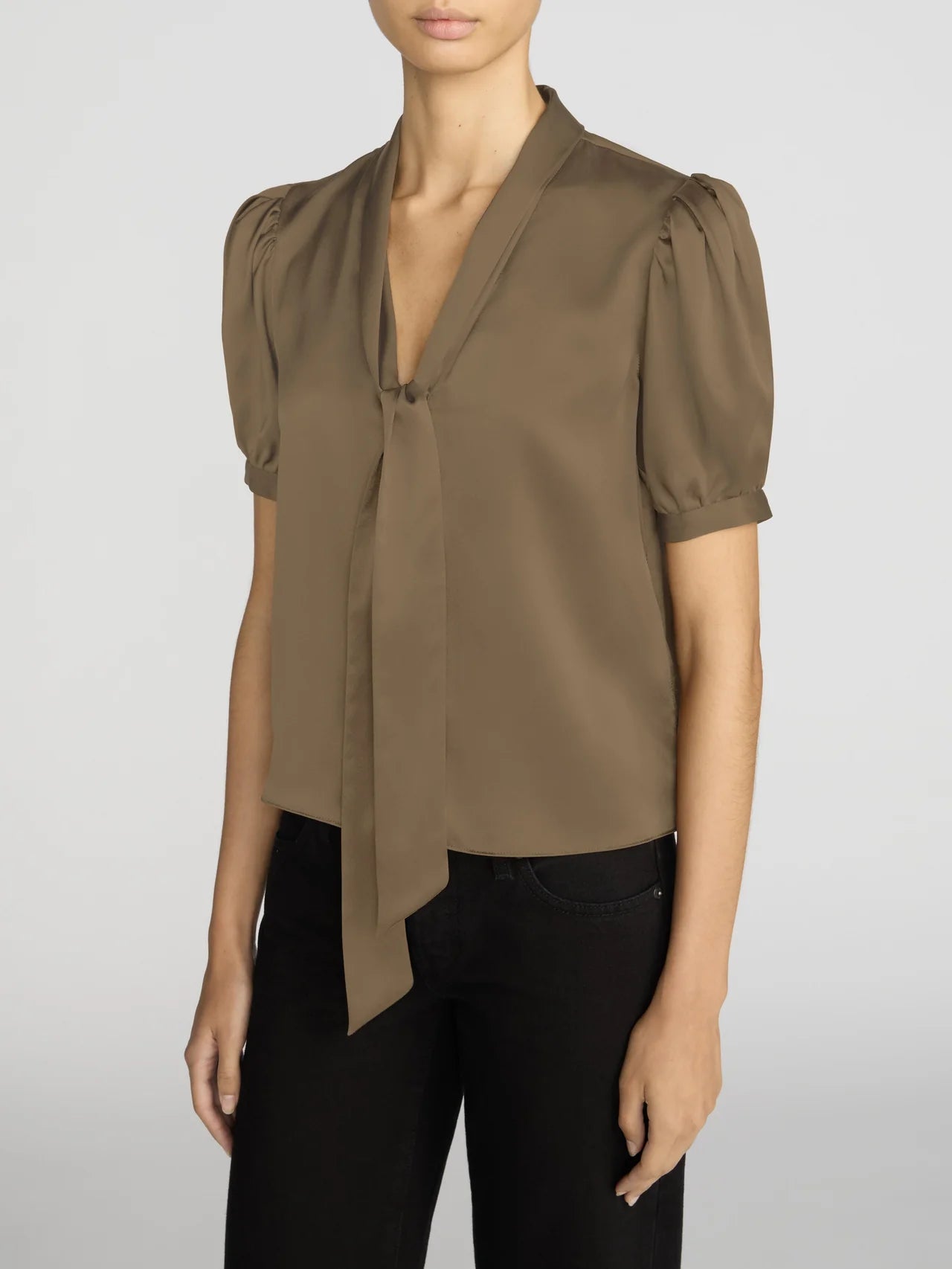 A woman wearing a taupe-brown short-sleeve blouse with puffed sleeves, a V-neckline, and a front tie detail, paired with black pants, against a light gray studio background.