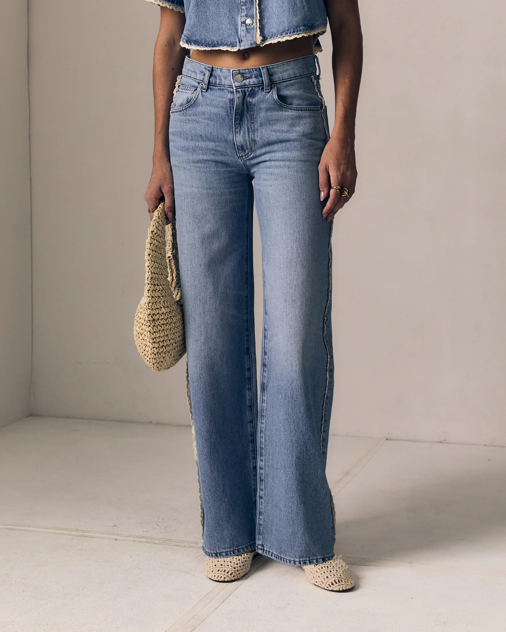 Person wearing blue jeans and a denim jacket holding a woven bag on a plain background