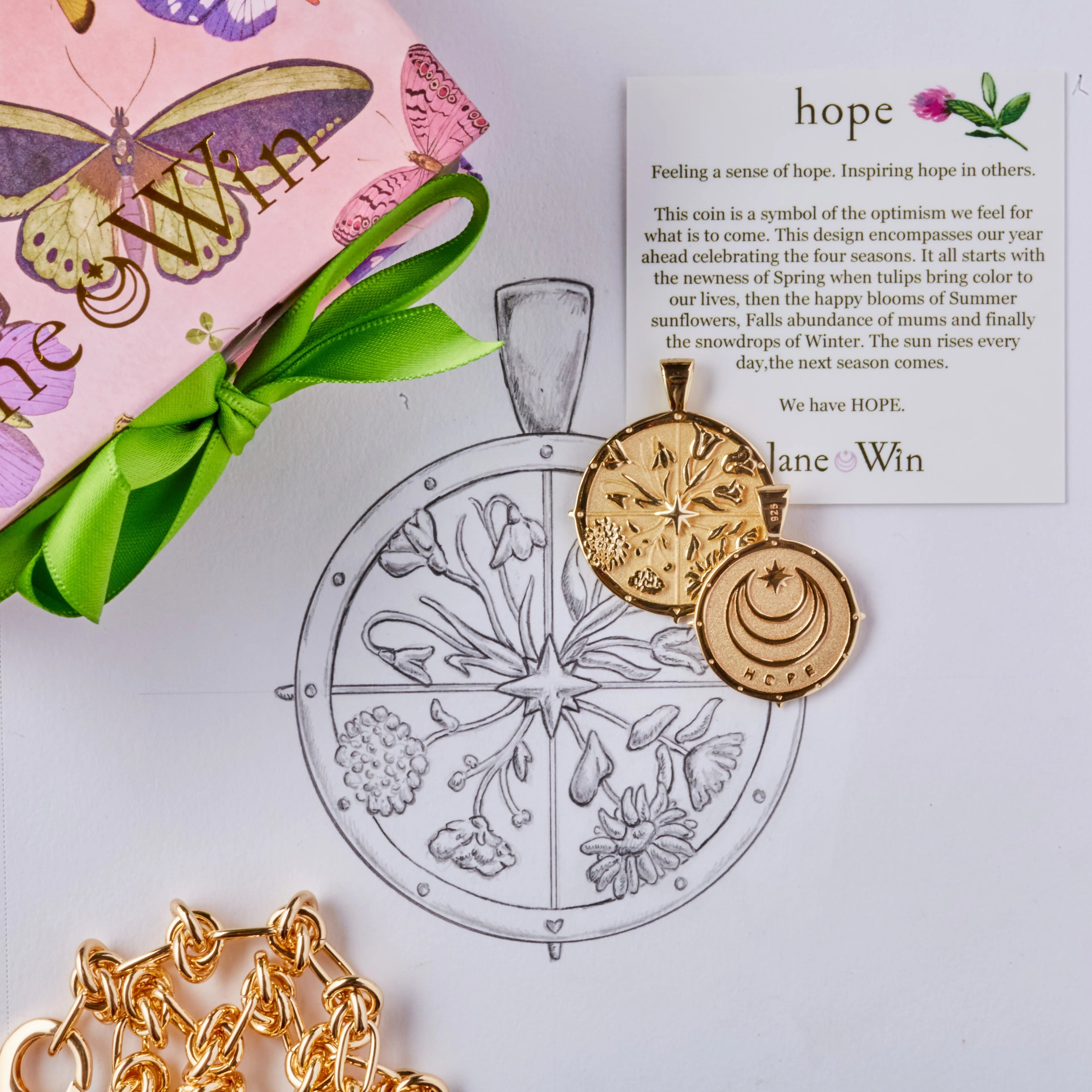 Gold pendant with chain, pink box with green ribbon, and 'hope' card on a light gray background