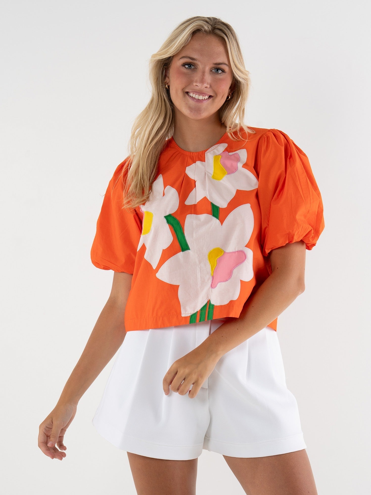 Woman wearing a bright orange top with floral designs and white shorts on a plain background