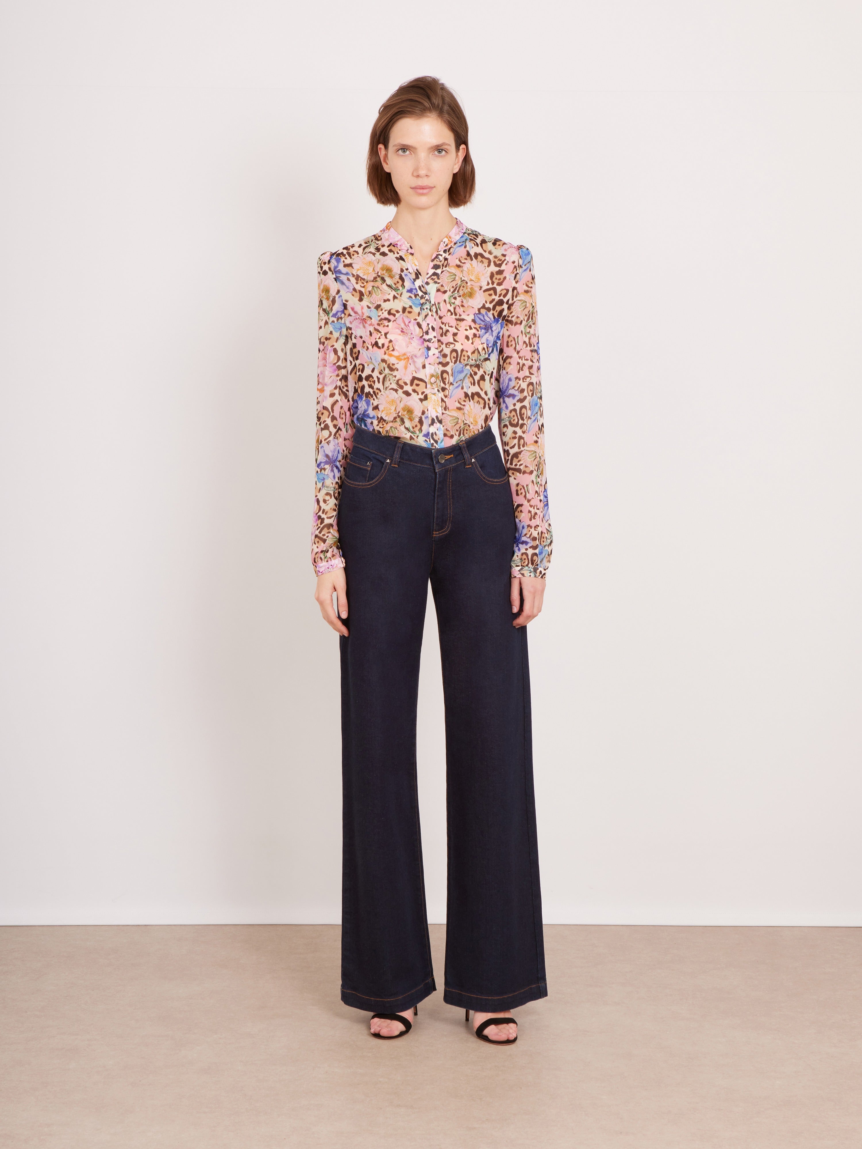 A model stands against a plain off-white wall wearing a long-sleeve button-up blouse with a dense multicolor floral and paisley print, paired with dark wide-leg jeans and black sandals.