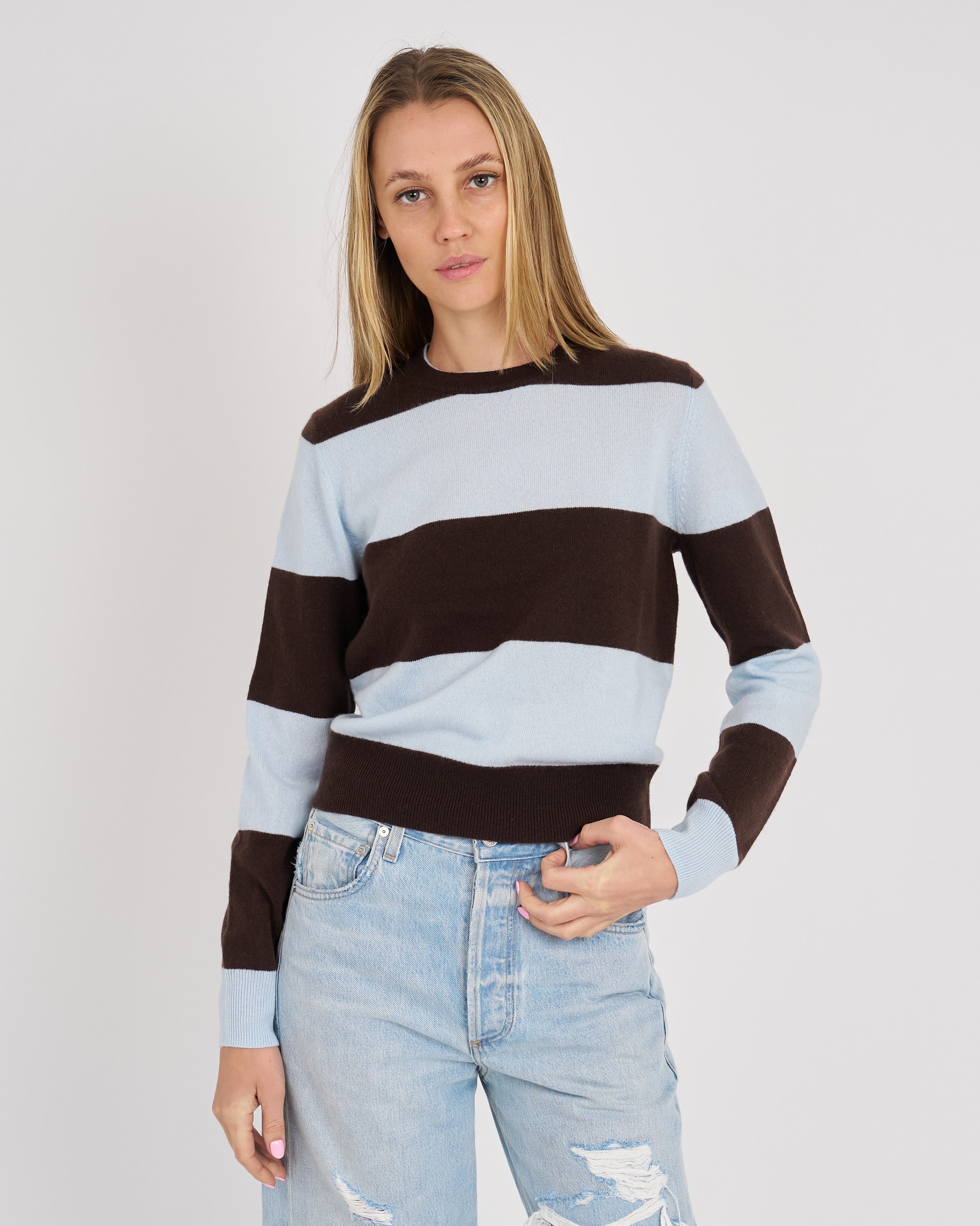 A woman wearing a brown and light blue horizontally striped knit sweater with light-wash distressed jeans, standing against a plain light gray studio background.