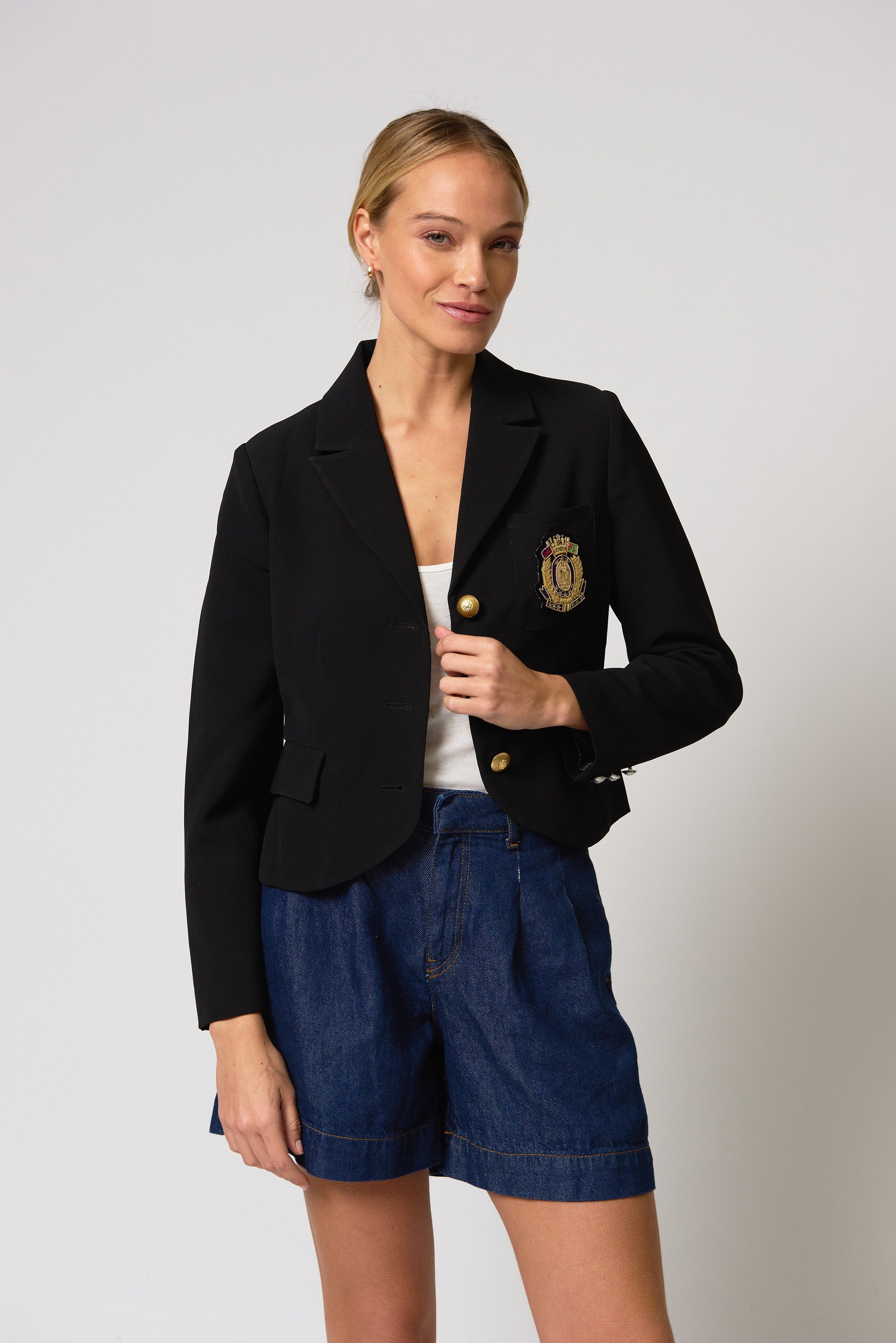 Woman wearing a black blazer with a logo and blue shorts on a light gray background