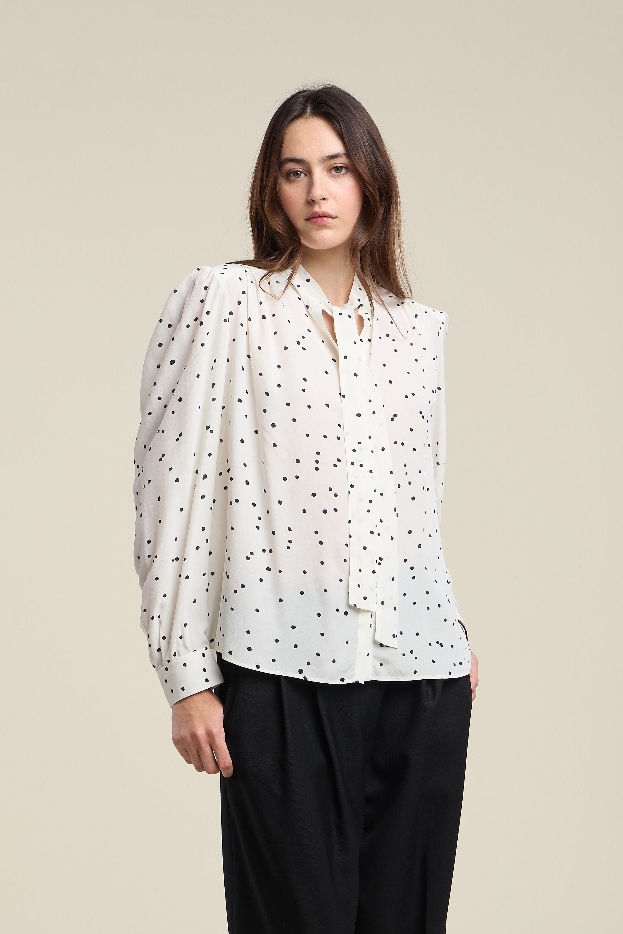 Woman wearing a white blouse with black polka dots and black pants on a beige background