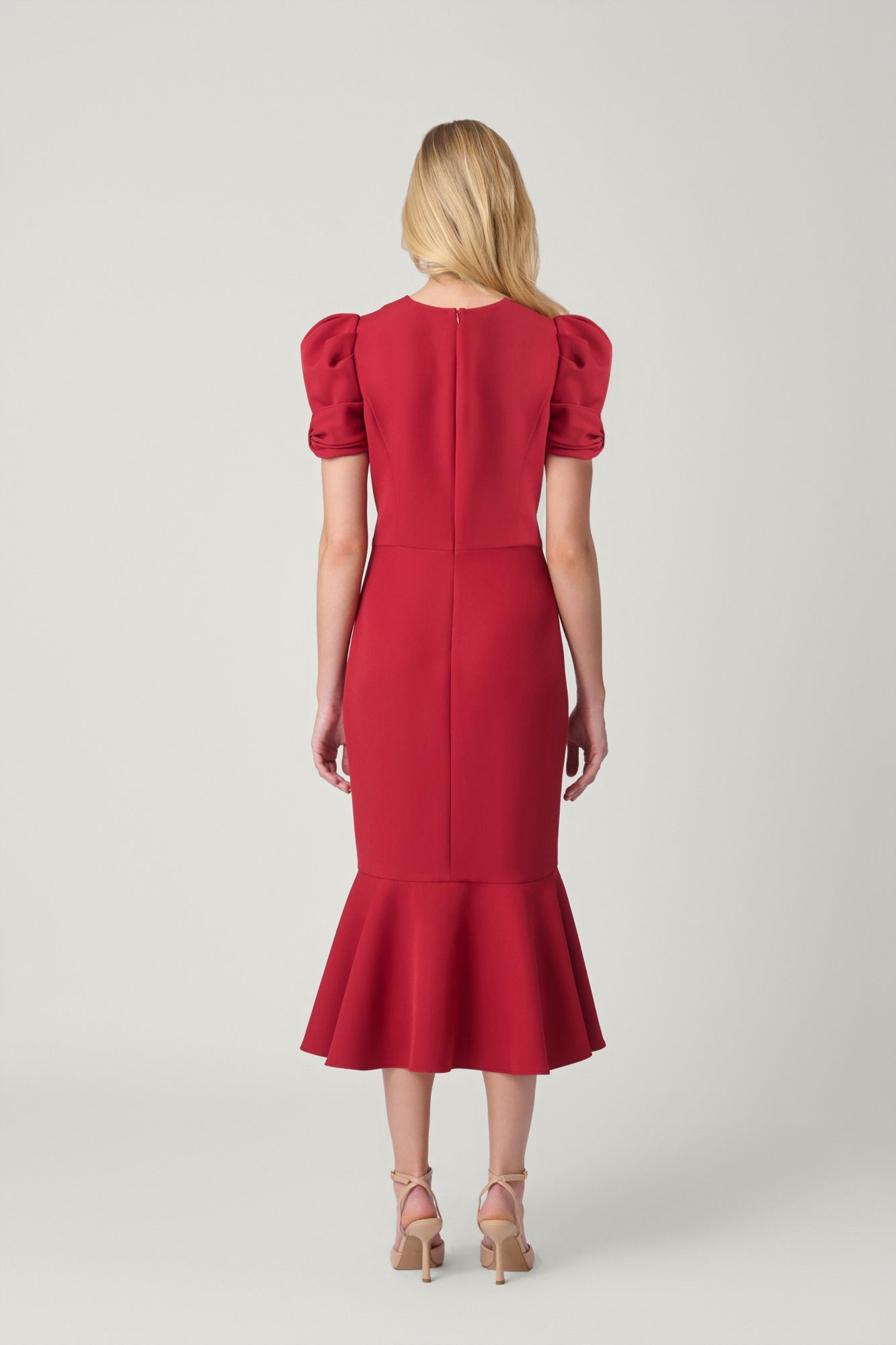 A woman viewed from the back wearing a bright red knee-length dress with short puff sleeves, a straight back seam, and a flared ruffle hem, paired with beige high-heeled sandals against a light gray studio background.