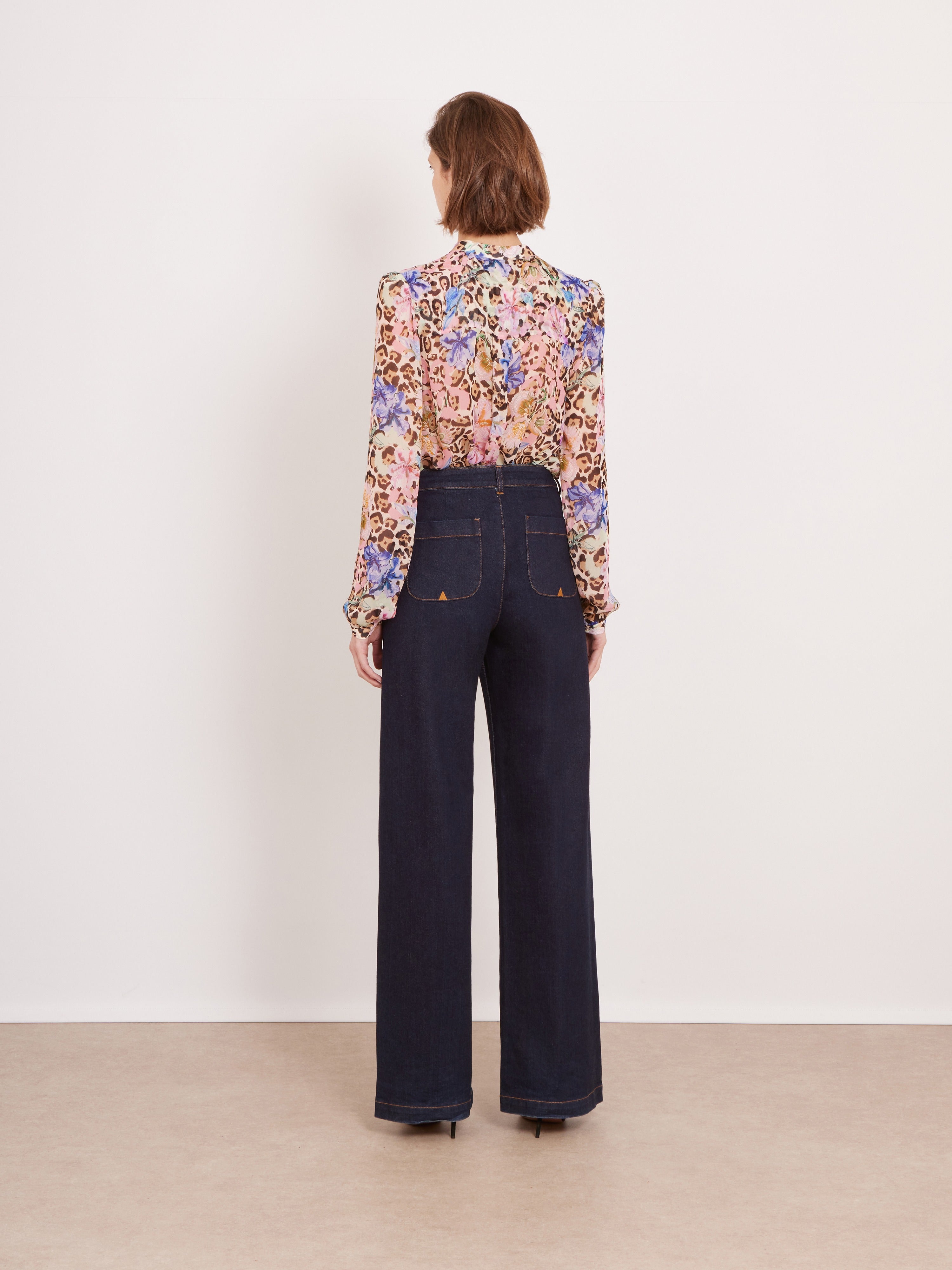 A model is shown from the back wearing a long-sleeve floral print blouse and dark wide-leg jeans against a plain off-white wall and beige floor.