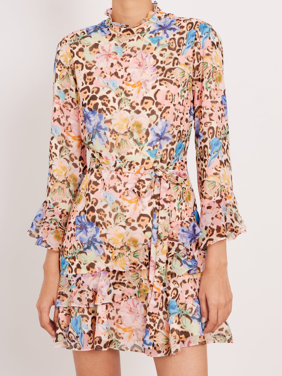 A model wearing a long-sleeve, ruffled mini dress with a busy leopard print and floral pattern in pink, blue, yellow, and brown on a light beige base, set against a plain light gray background.