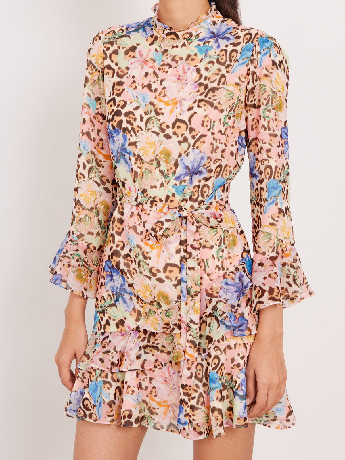A long-sleeve mini dress with a multicolor floral and leopard print pattern, featuring a high ruffled collar, a self-tie waist belt, and flared bell sleeves, shown on a model against a plain light background.