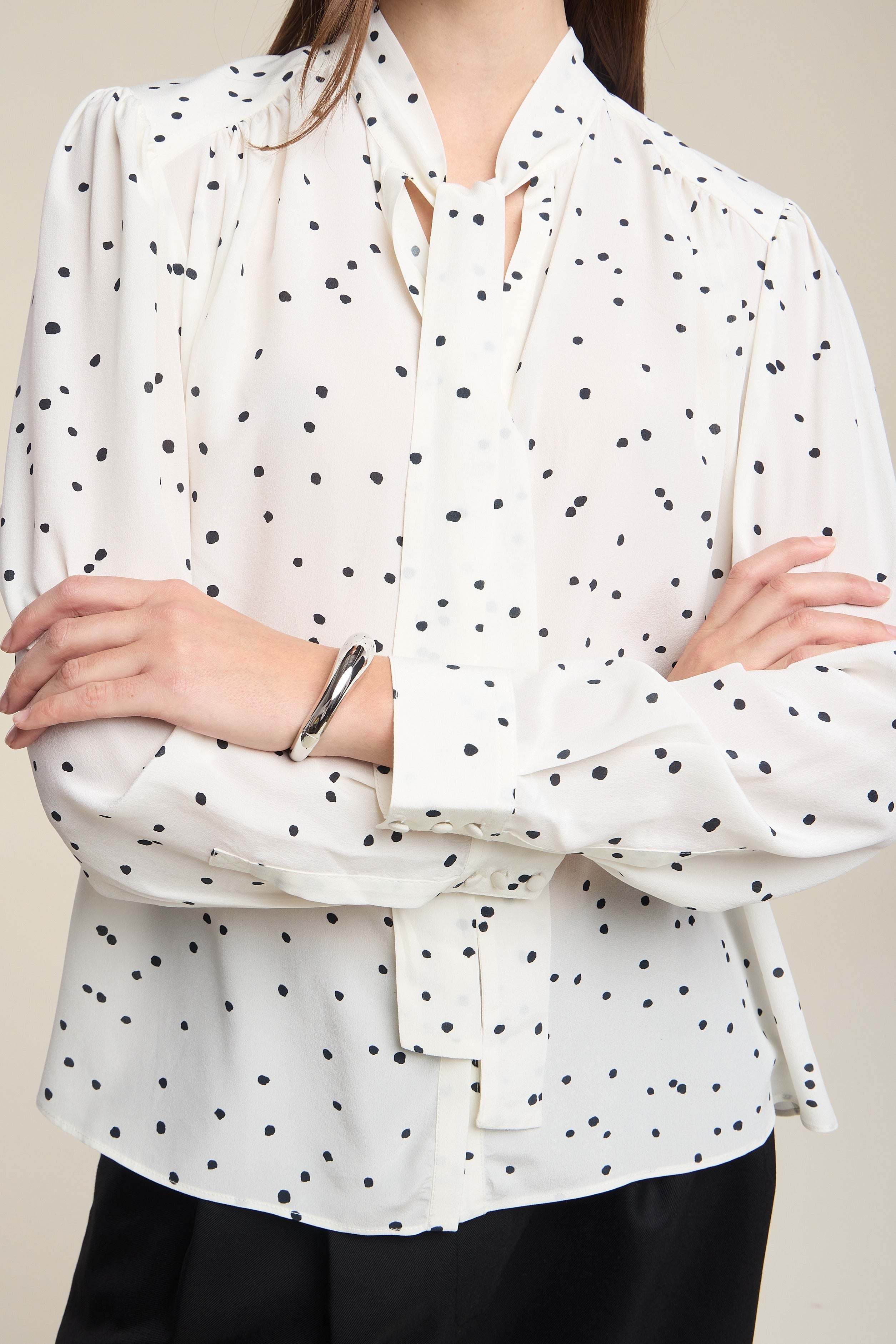 A woman wearing a white long-sleeve blouse with a black polka dot pattern, featuring a split neckline with tie details and a self-tie waist belt, paired with black pants.