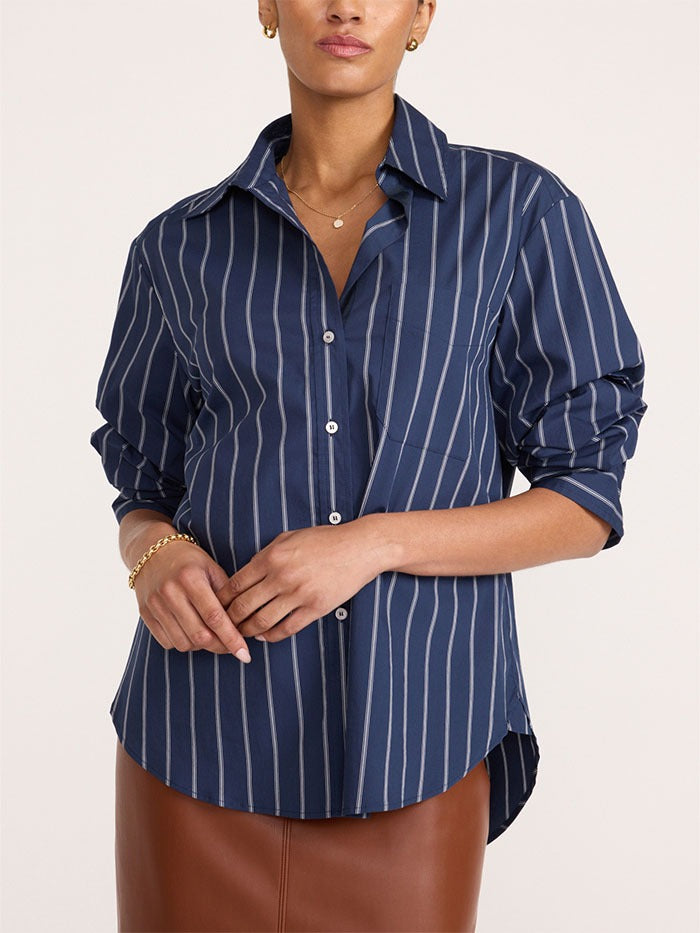 A woman wearing a navy blue button-up shirt with thin white vertical stripes, paired with a brown leather skirt, against a plain light beige background.