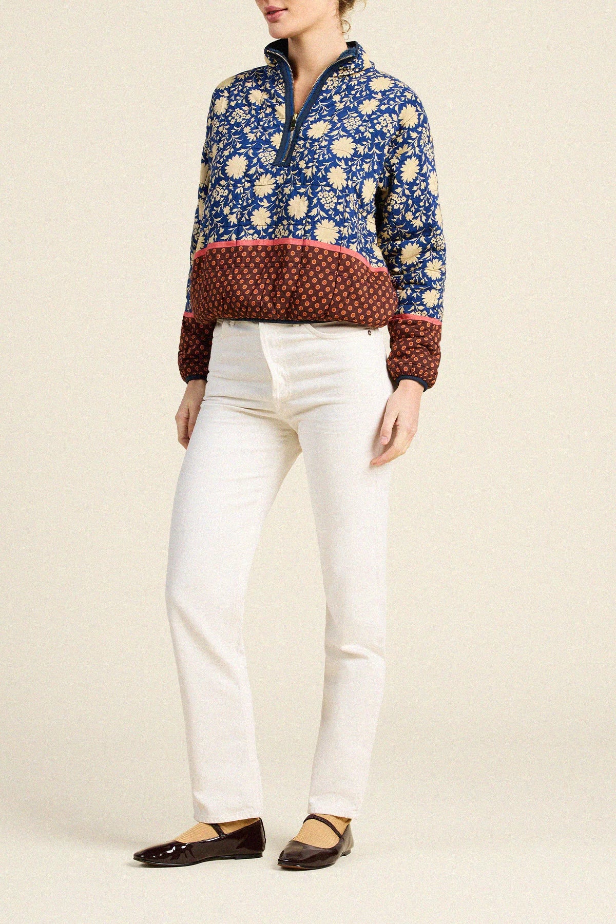 A model wearing a blue and beige floral quarter-zip pullover with a burgundy patterned hem, paired with white pants and dark brown flats, against a solid light beige background.