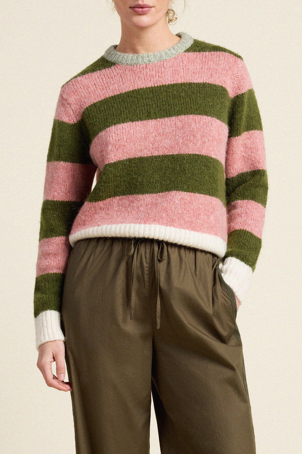 A woman wearing a fuzzy knit sweater with horizontal stripes in olive green, dusty pink, and off-white, paired with olive green drawstring trousers, against a plain beige background.