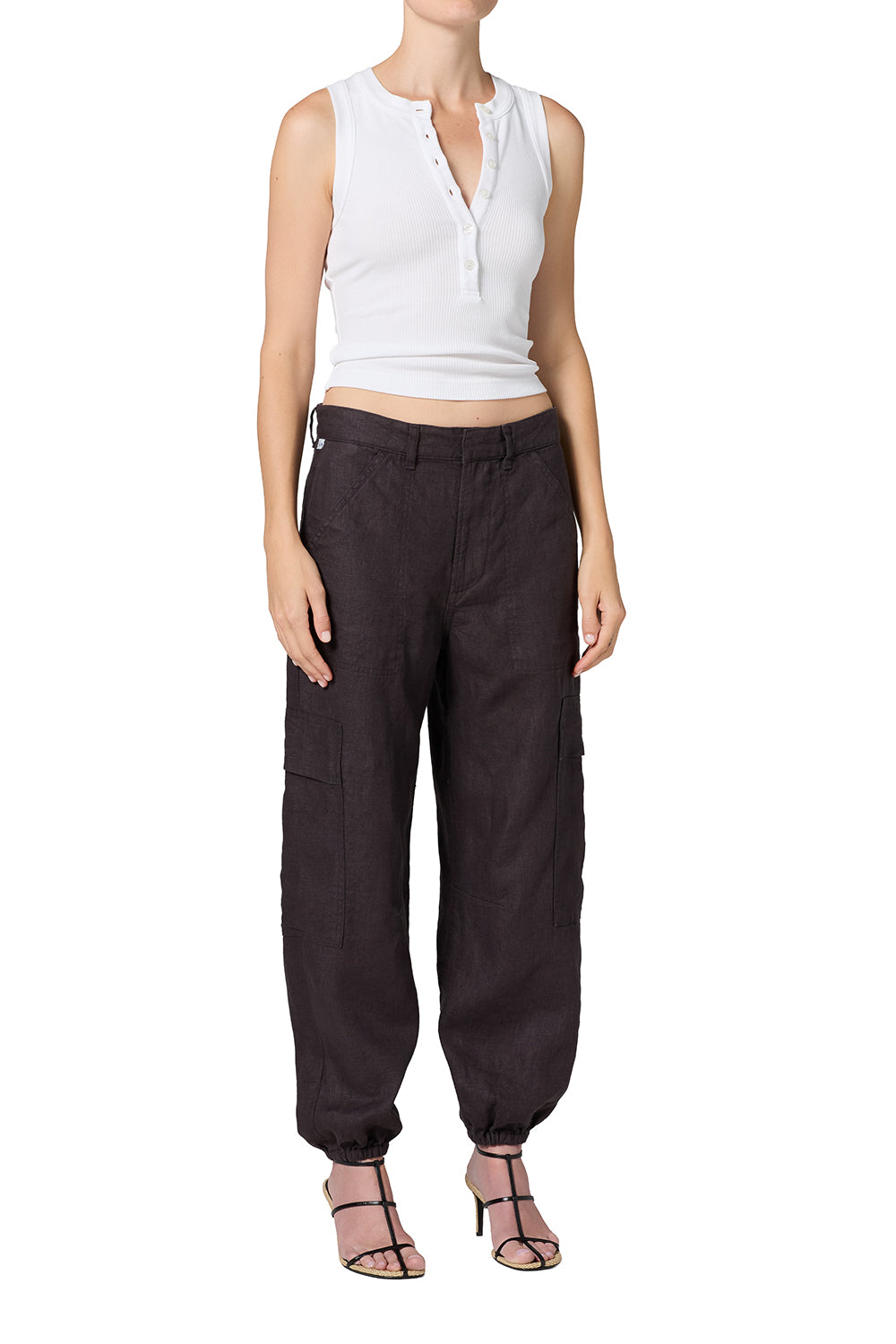 A model wearing dark brown baggy cargo pants with a white sleeveless crop top and strappy heels, shown against a plain white background.