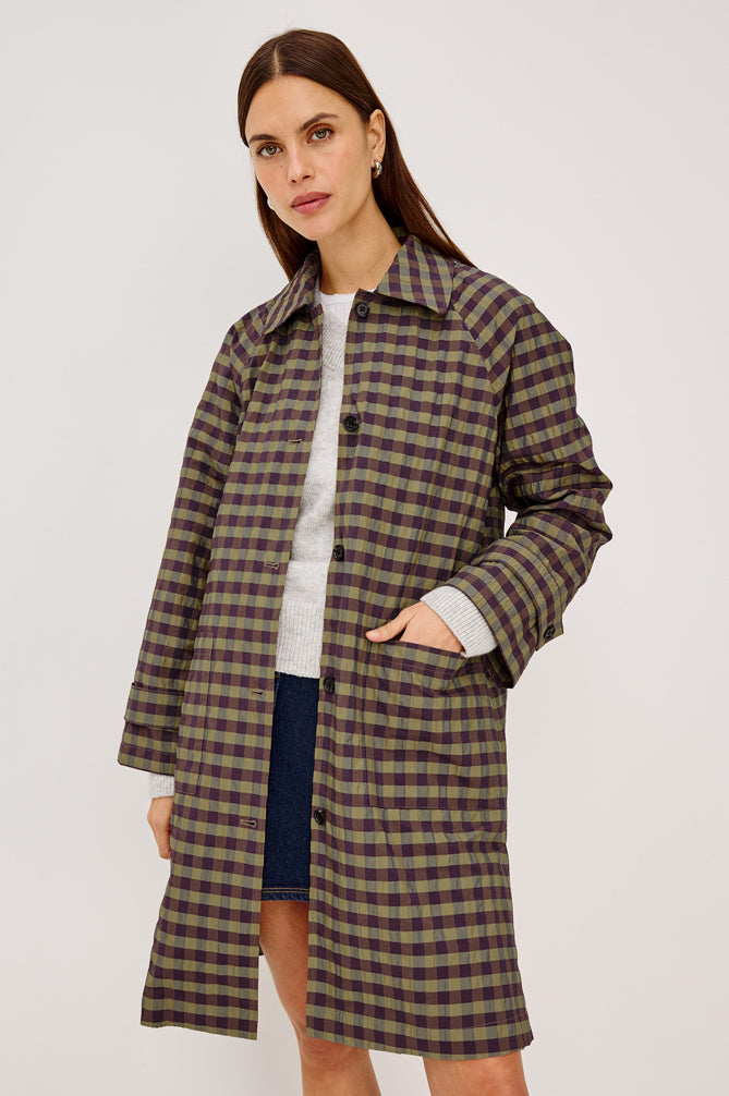 A woman modeling a mid-length plaid coat in brown and olive green tones over a white top and navy skirt, photographed against a plain light gray studio background.