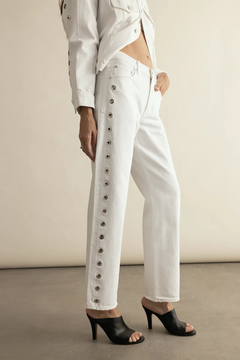 Woman wearing a white denim outfit with studded details against a neutral background and heeled mules.