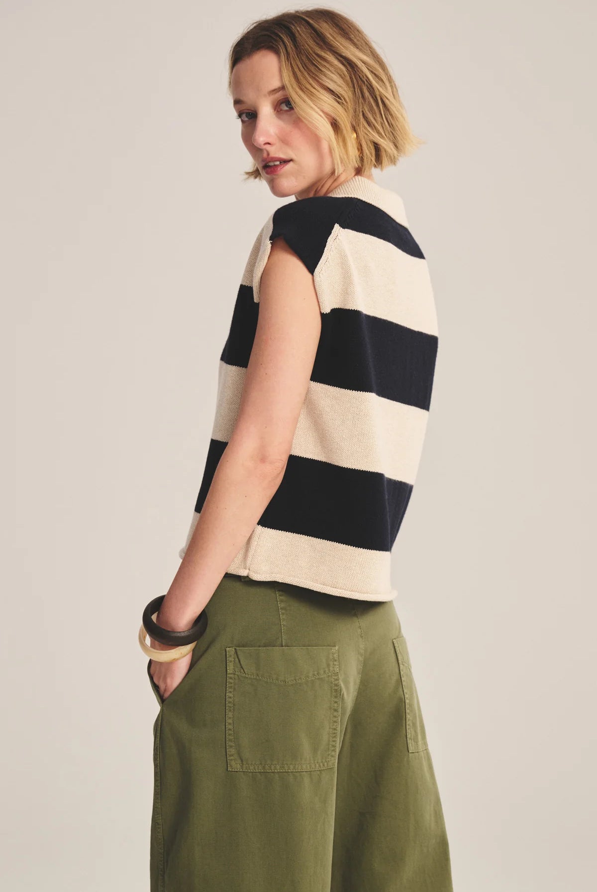 Woman wearing a black and white striped sleeveless top with green pants on a plain background