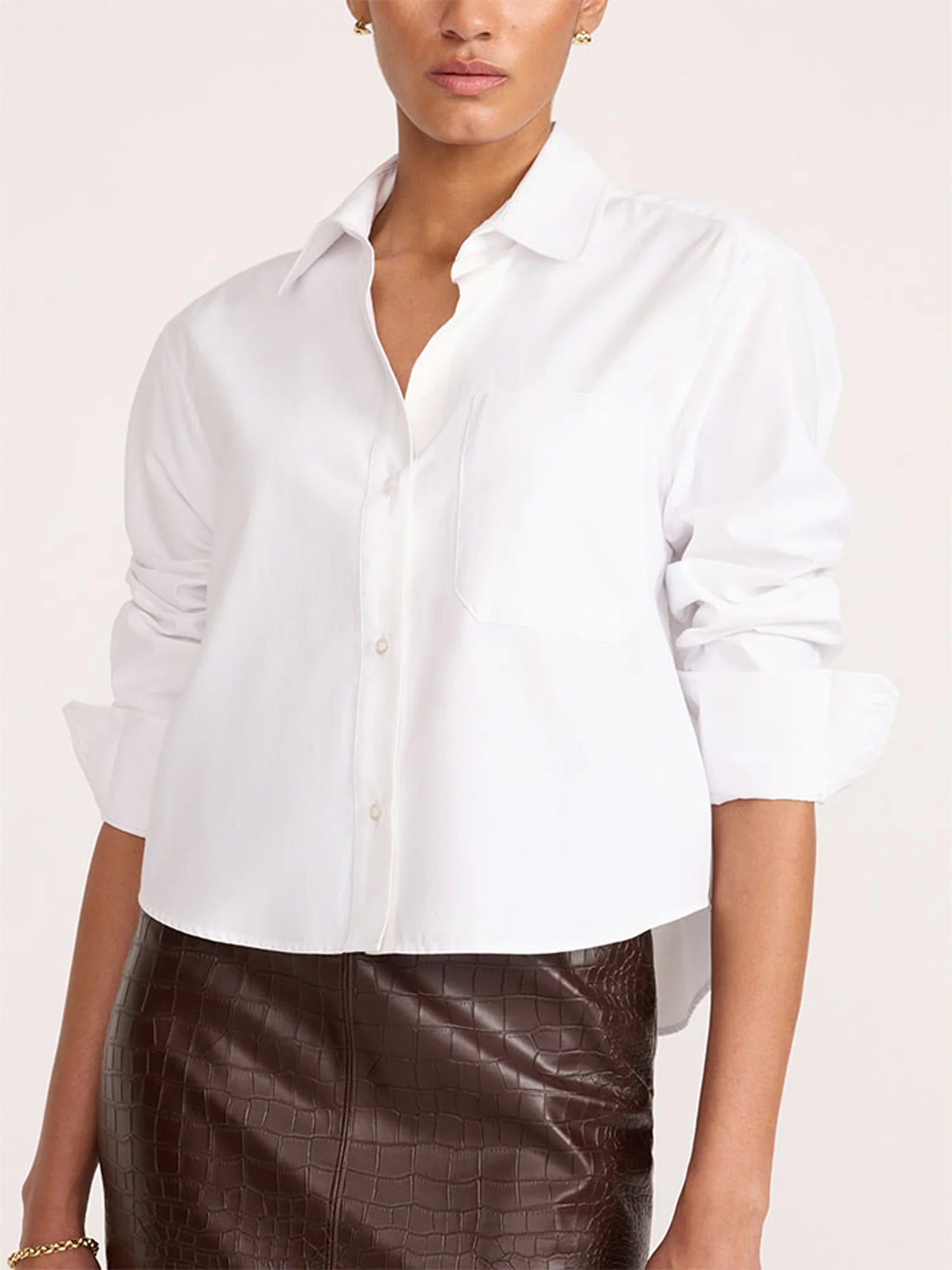 A woman wearing a white cropped button-up shirt with rolled sleeves and a brown textured skirt, photographed against a plain light beige studio background.
