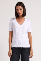 Woman wearing a white cable knit sweater with black pants on a plain background
