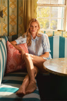 Woman sitting on a striped sofa in a sunlit room with floral wallpaper.