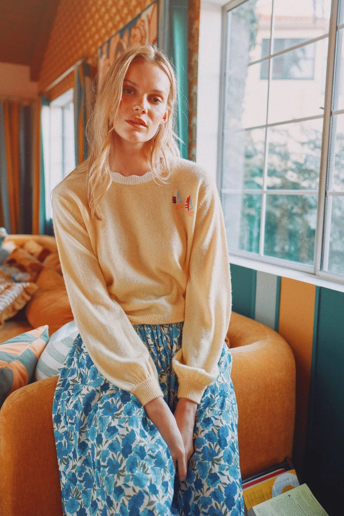 Woman wearing a beige sweater and blue floral pants in a room with orange chairs and large windows.