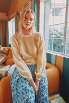 Woman wearing a beige sweater and blue floral pants in a room with orange chairs and large windows.