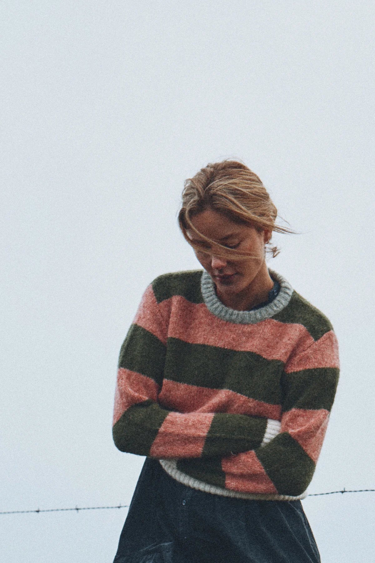 A woman wearing a fuzzy knit sweater with horizontal stripes in dusty pink, dark green, and gray, paired with a dark skirt, set against a pale gray sky.