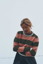A woman wearing a fuzzy knit sweater with horizontal stripes in dusty pink, dark green, and gray, paired with a dark skirt, set against a pale gray sky.