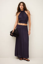 Woman wearing a dark purple outfit with a long skirt and top against a plain background
