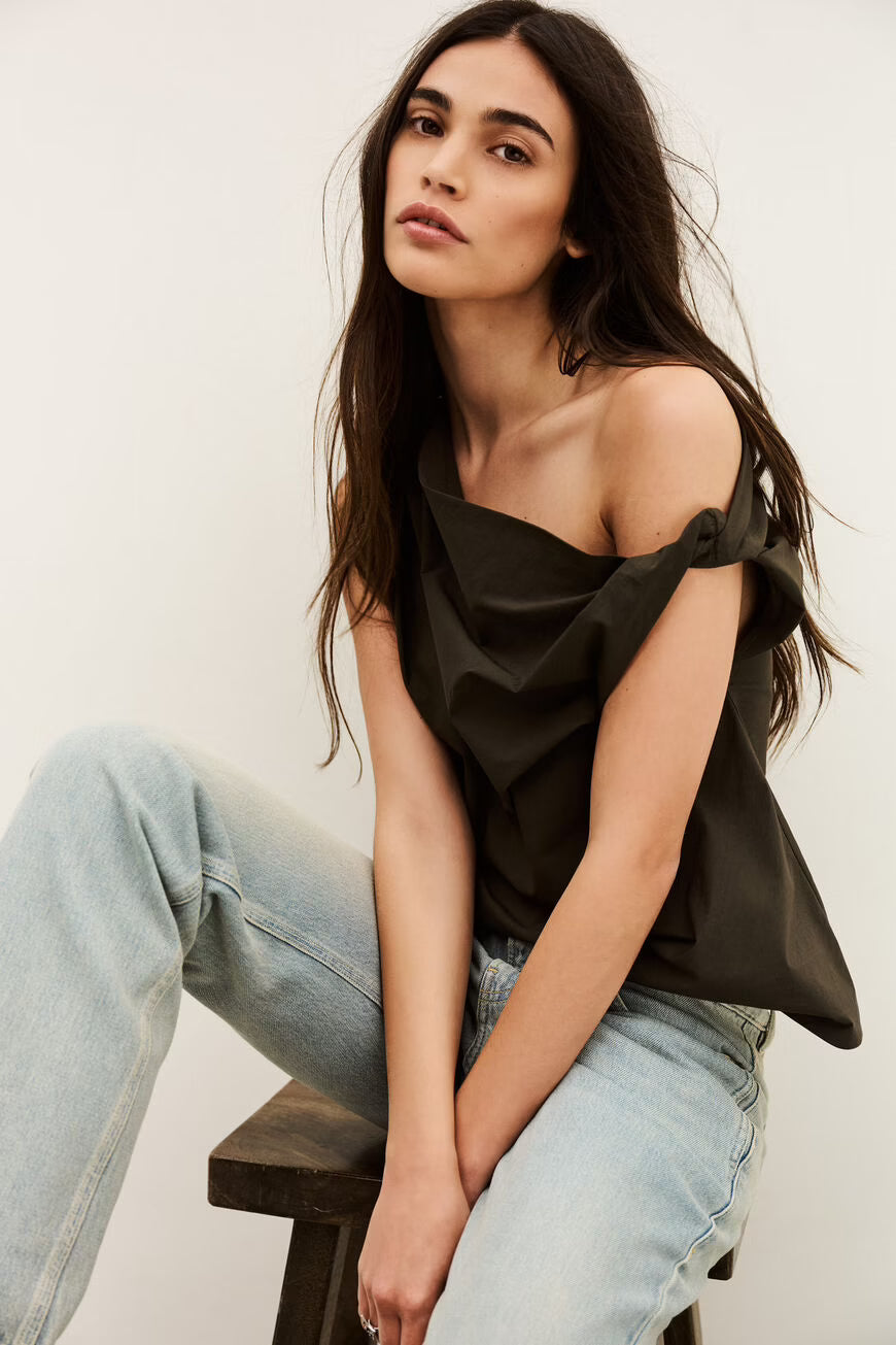 Woman wearing a black off-shoulder top and light blue jeans sitting on a wooden stool.