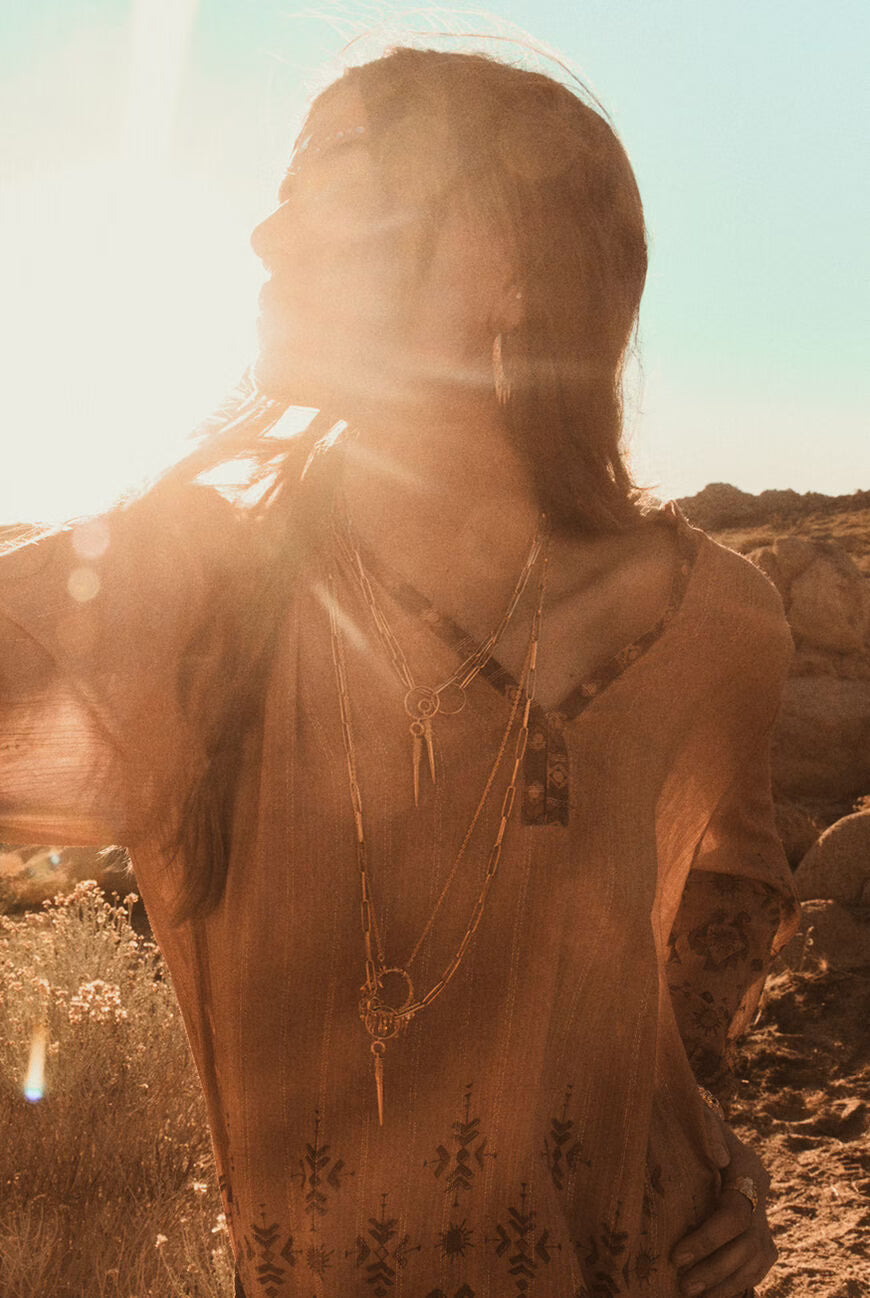 Woman with layered necklaces standing in a desert landscape with sunlight filtering through.