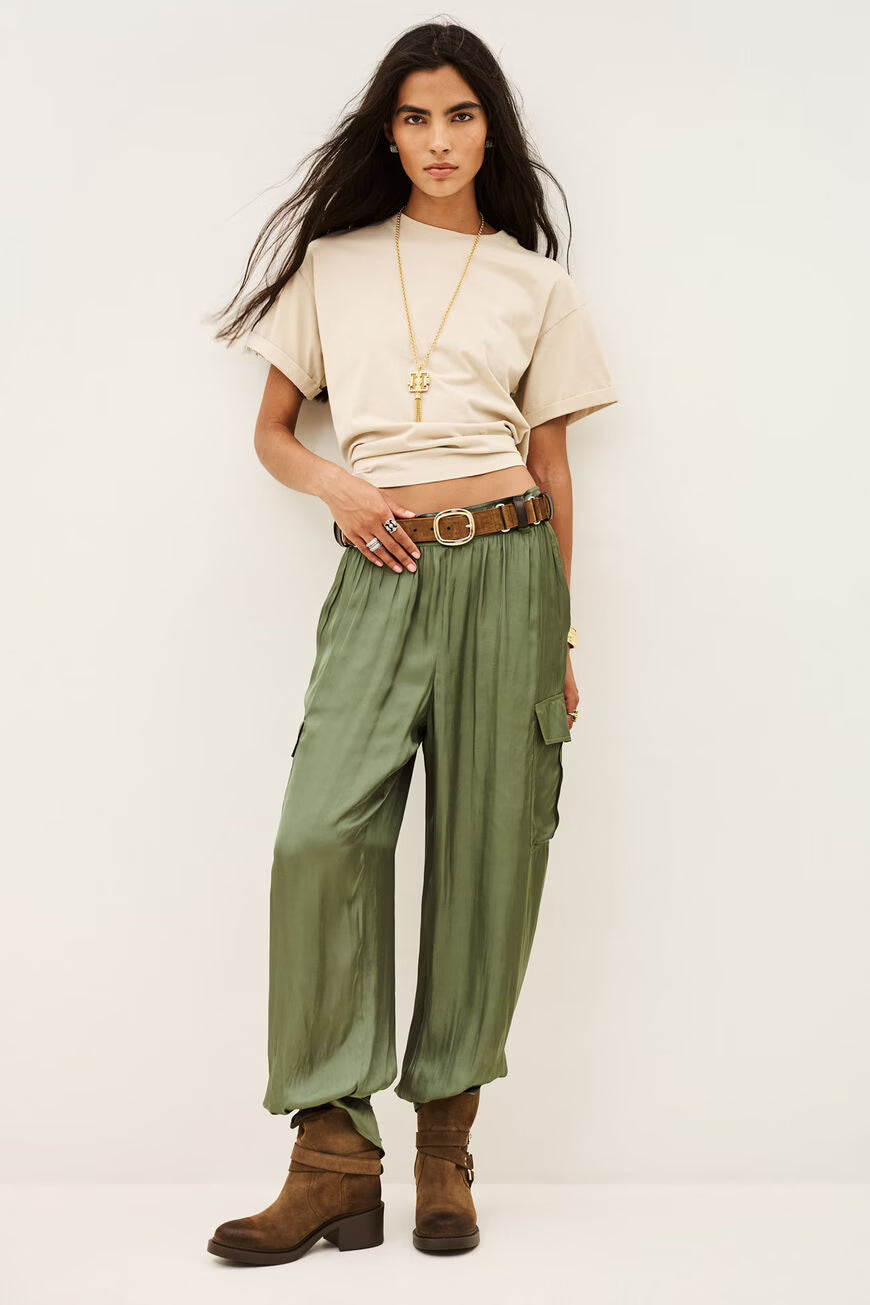 A model stands against a plain off-white studio background wearing a beige short-sleeve t-shirt, olive green wide-leg cargo pants, a brown leather belt with a gold buckle, and brown suede boots.