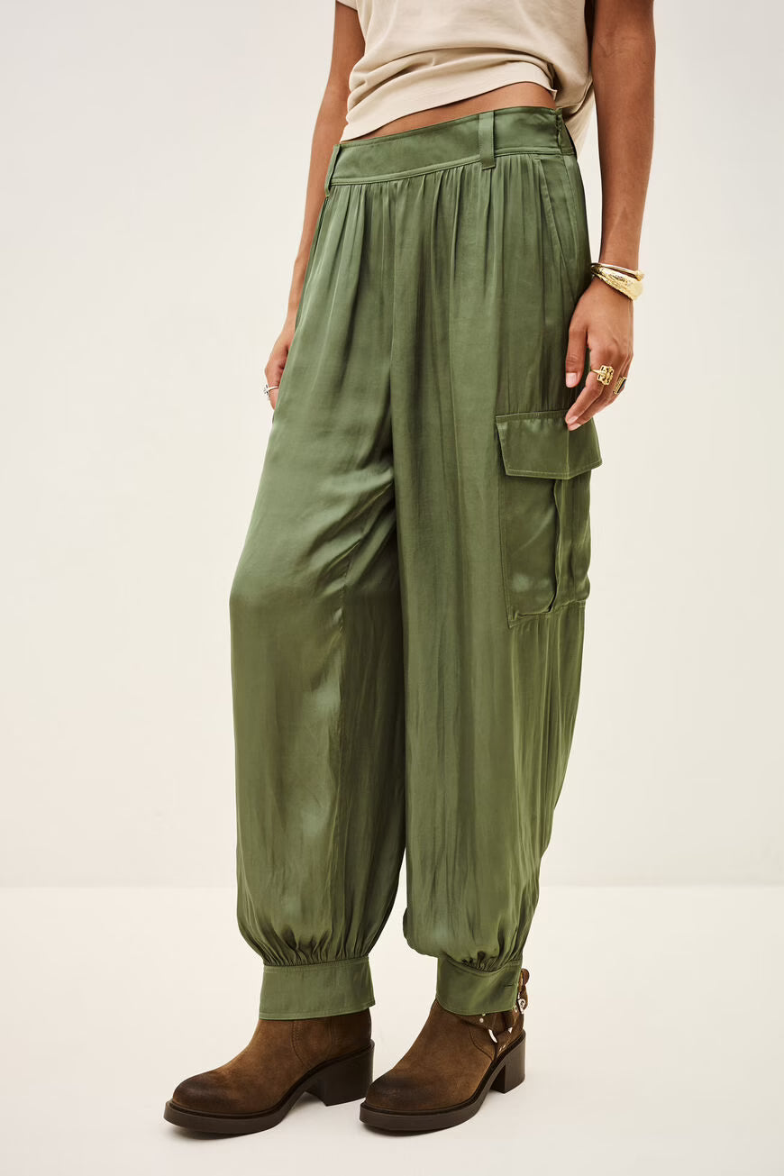 A person wearing olive green harem-style cargo pants with a beige cropped top and brown ankle boots, photographed against a plain light studio background.