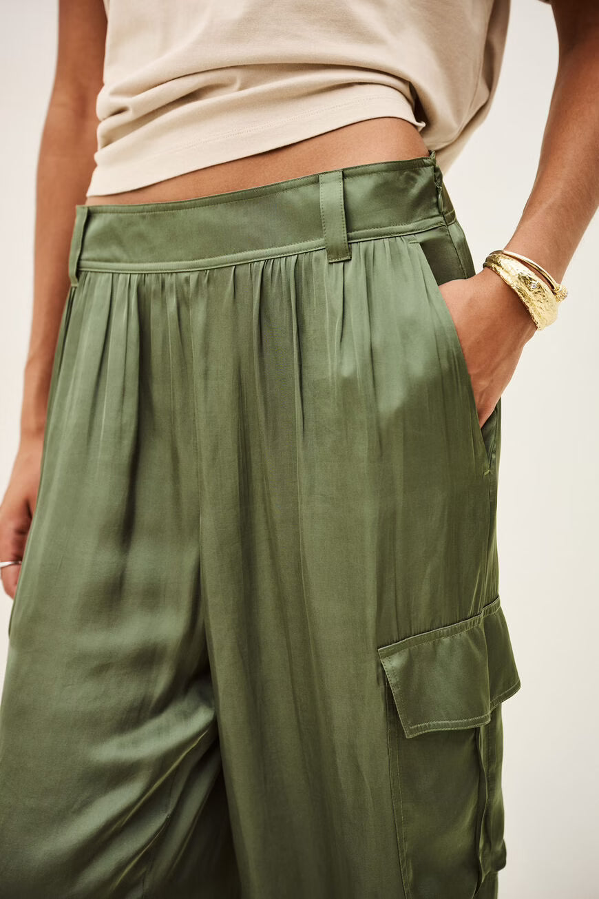 Close-up of a person wearing olive green wide-leg cargo pants with a satin-like sheen, paired with a beige cropped top. The model has a gold bracelet on one wrist, and the background is a clean off-white studio setting.