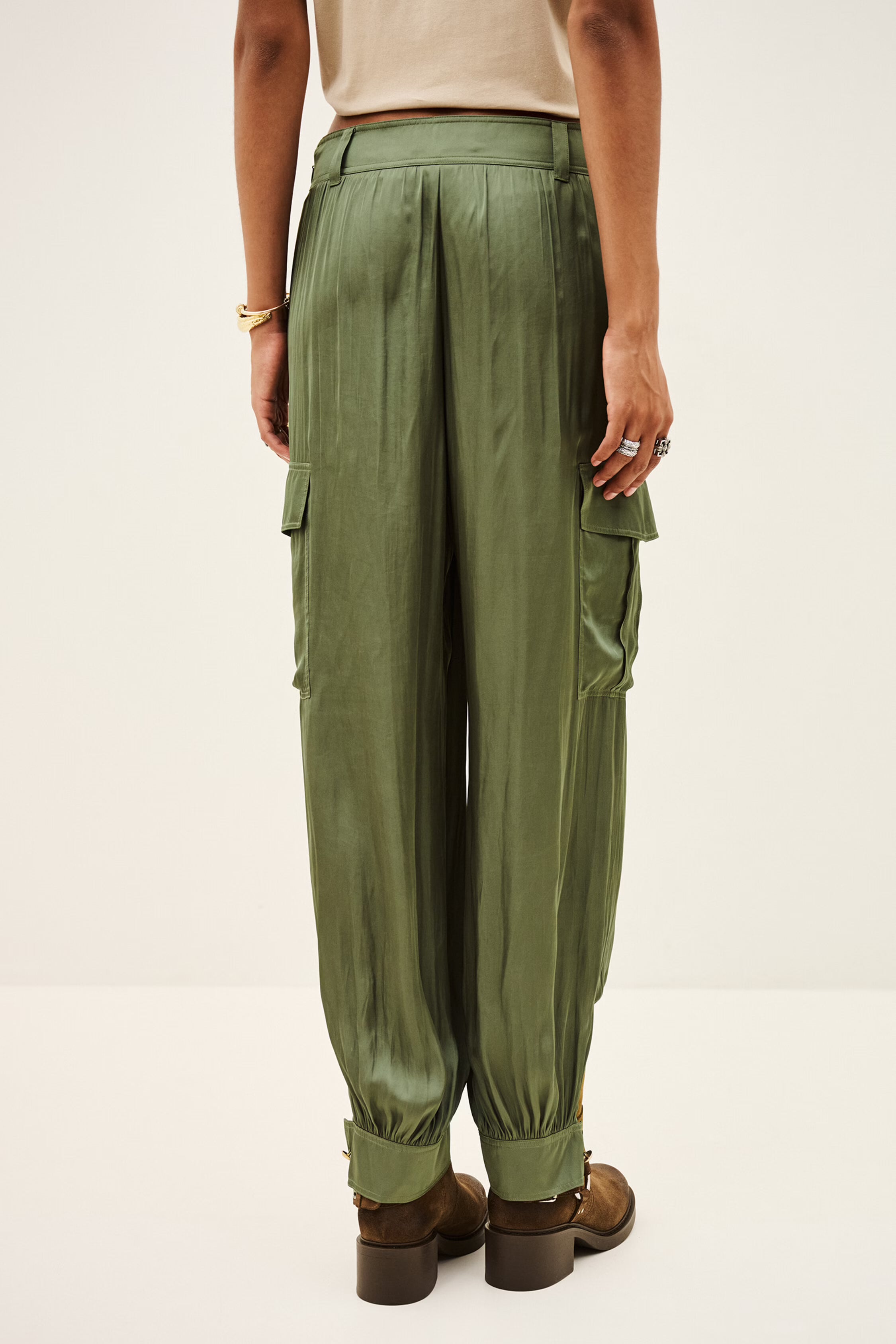 A person shown from the back wearing olive green cargo pants with side flap pockets, a light beige top, and brown shoes against a plain light background.