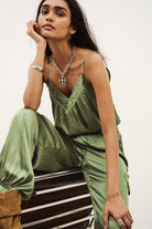 A woman models an olive green satin jumpsuit with matching wide-leg pants, styled with silver jewelry, sitting on a striped wooden bench against a plain white background.