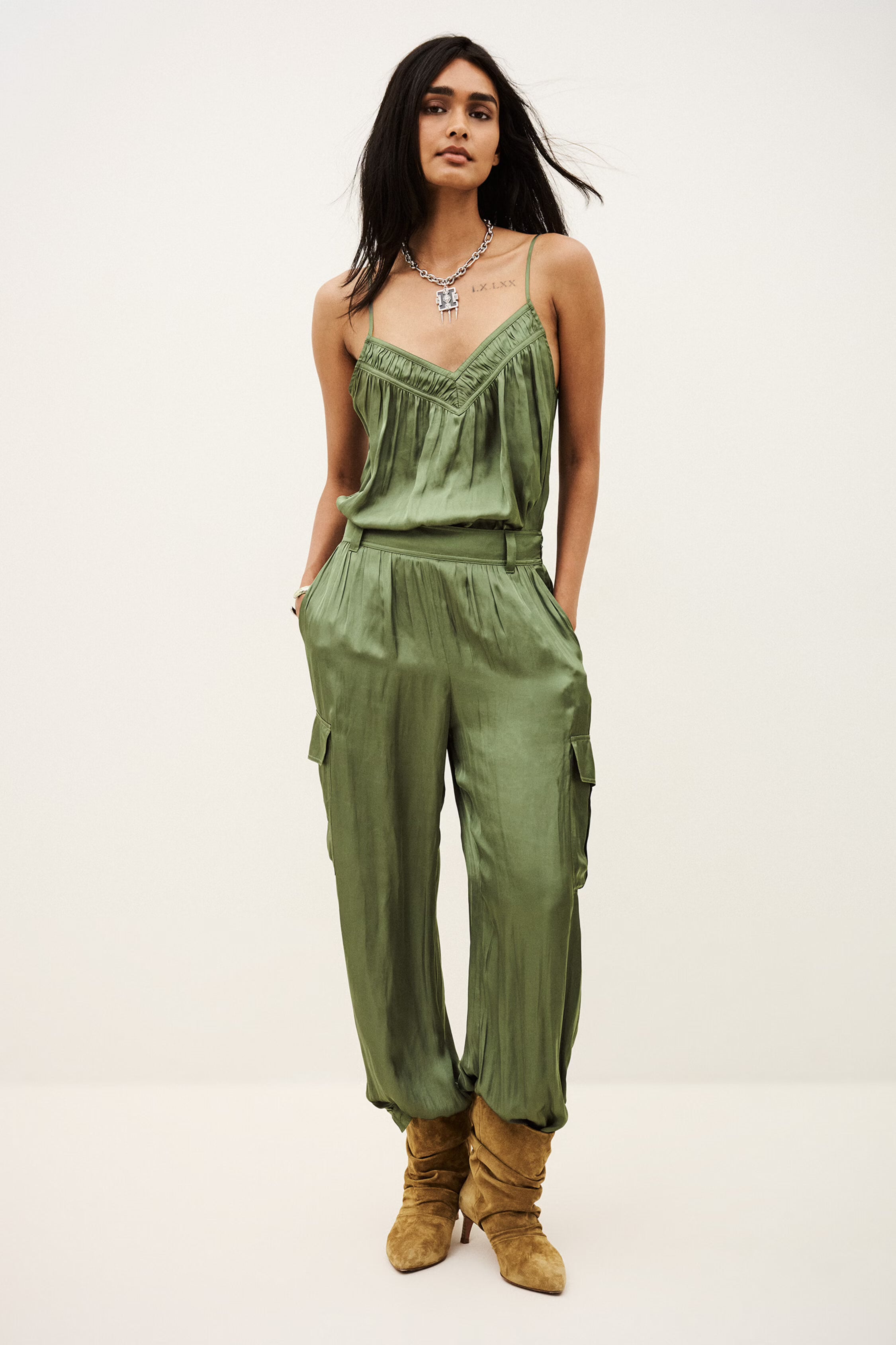 A woman modeling a green satin jumpsuit with a ruffled sweetheart neckline and side cargo pockets, paired with tan suede boots, against a plain white studio background.