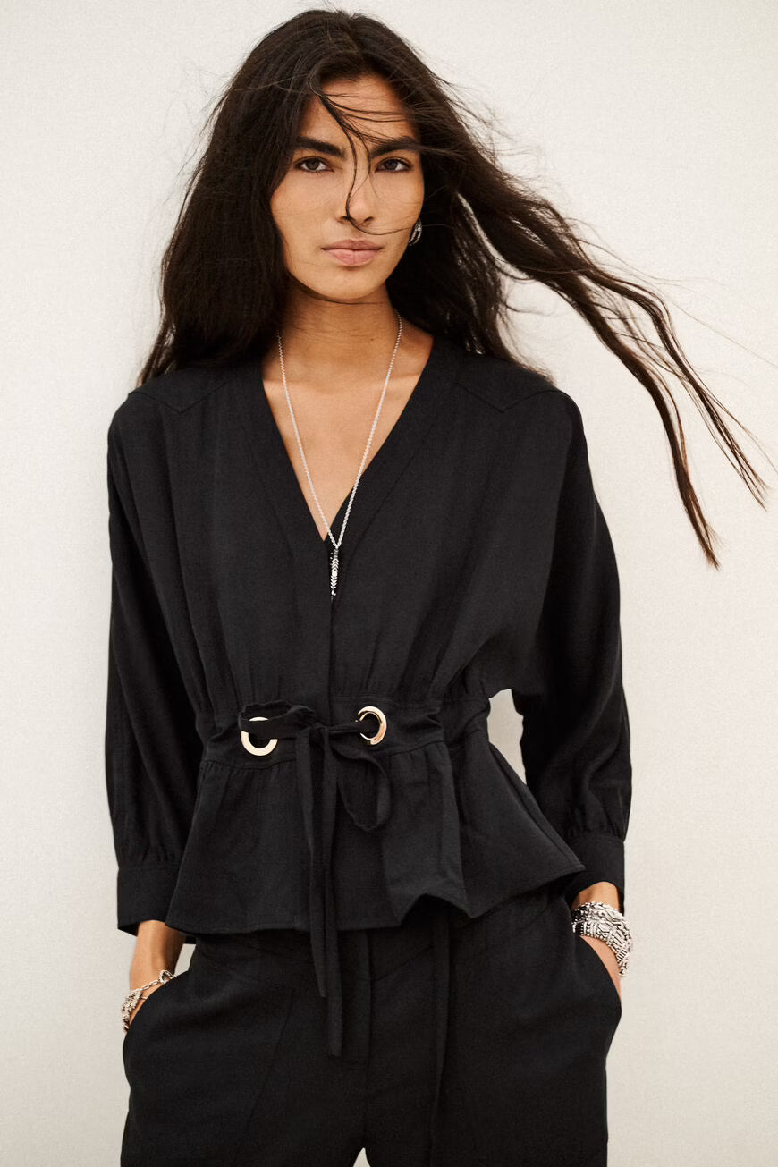 A woman modeling a black long-sleeve belted top with a peplum hem and drawstring waist, paired with black pants, against a plain light background.